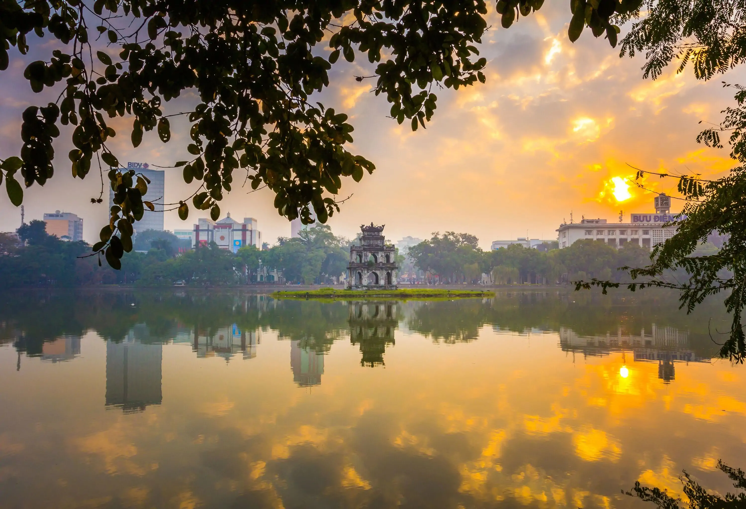 A peaceful lake with a small tower in the middle reflects the nearby structures and the setting sun.