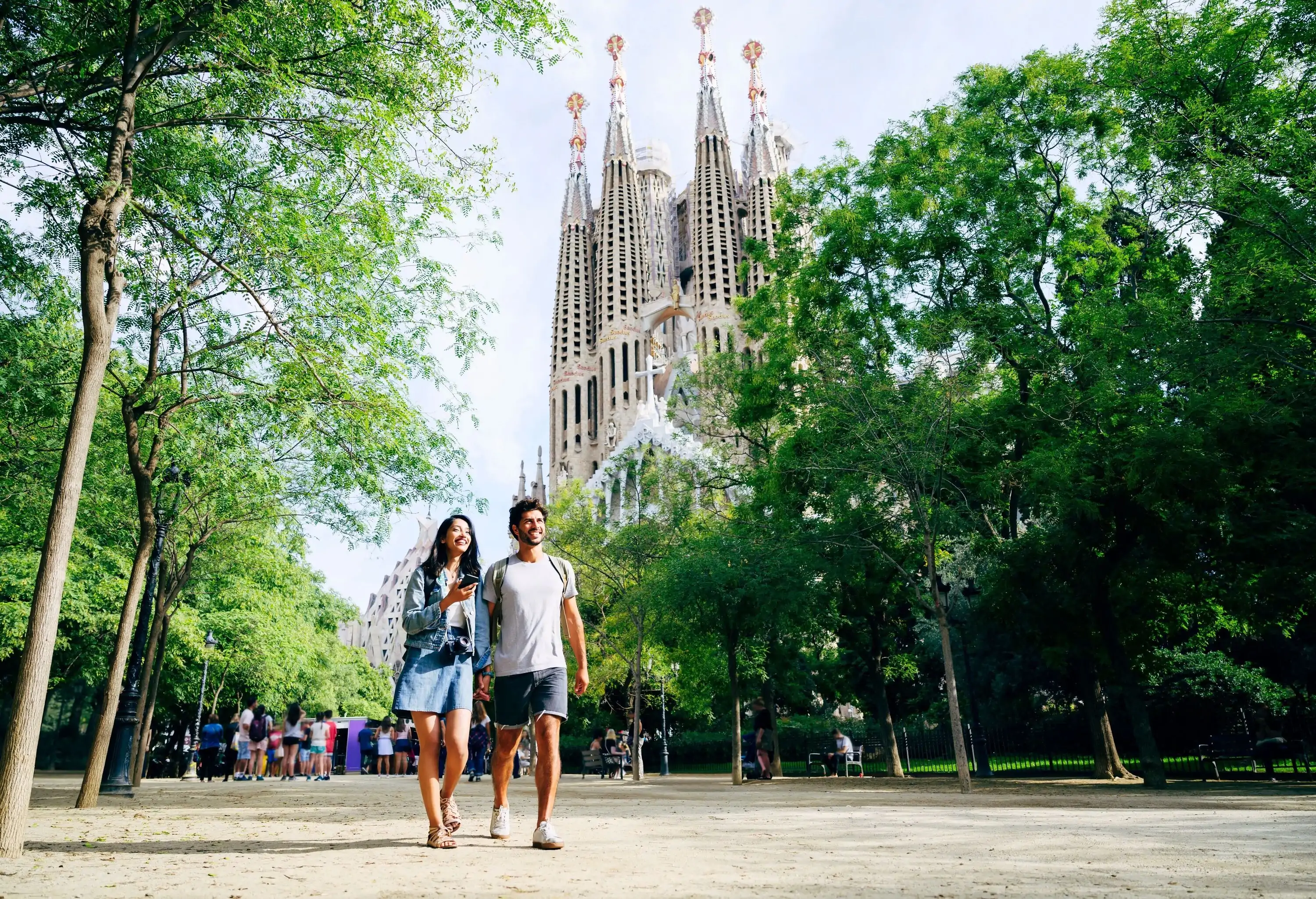 A couple strolling in the park with the Sagrada Familia emerging through the trees in the background.