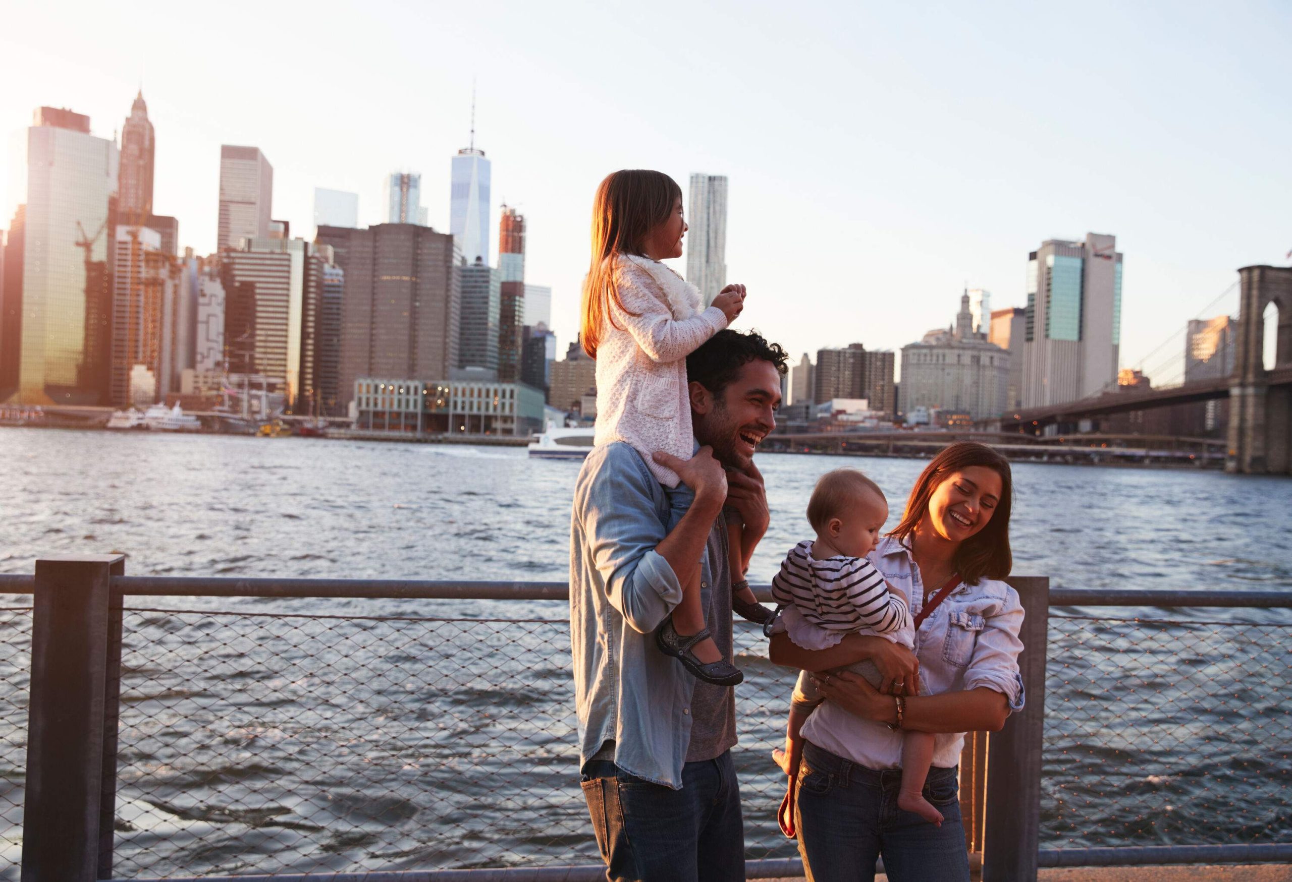 A young couple standing by the river with their two children, with skyscrapers in the backdrop.