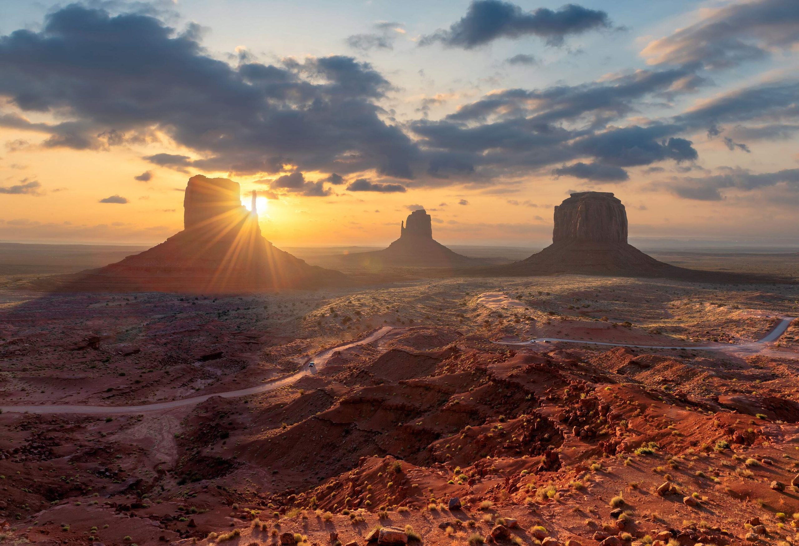 The sun rises in a dramatic sky above three iconic rock formations emerging in the rugged valley.