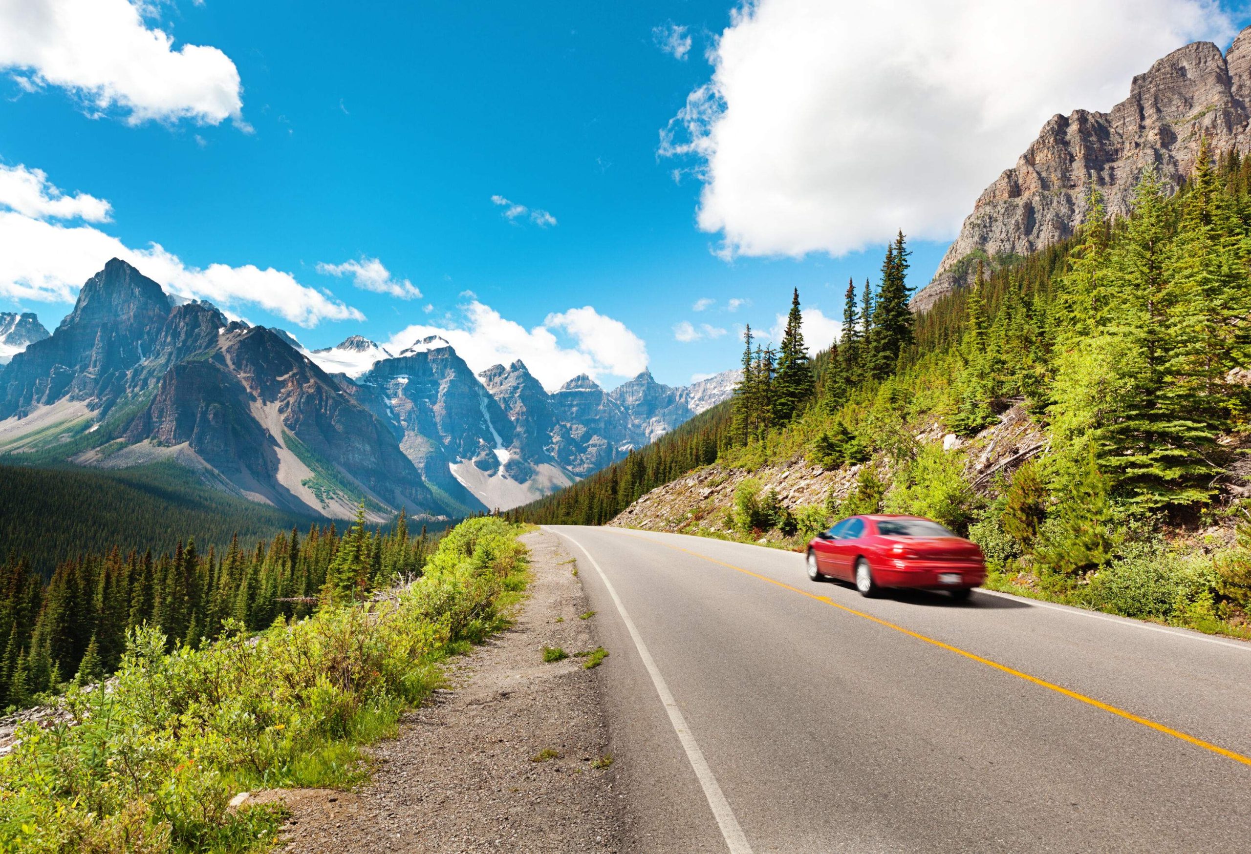 A lone red car driving towards the imposing rock mountain ranges with pointed peaks on a bright sunny day.