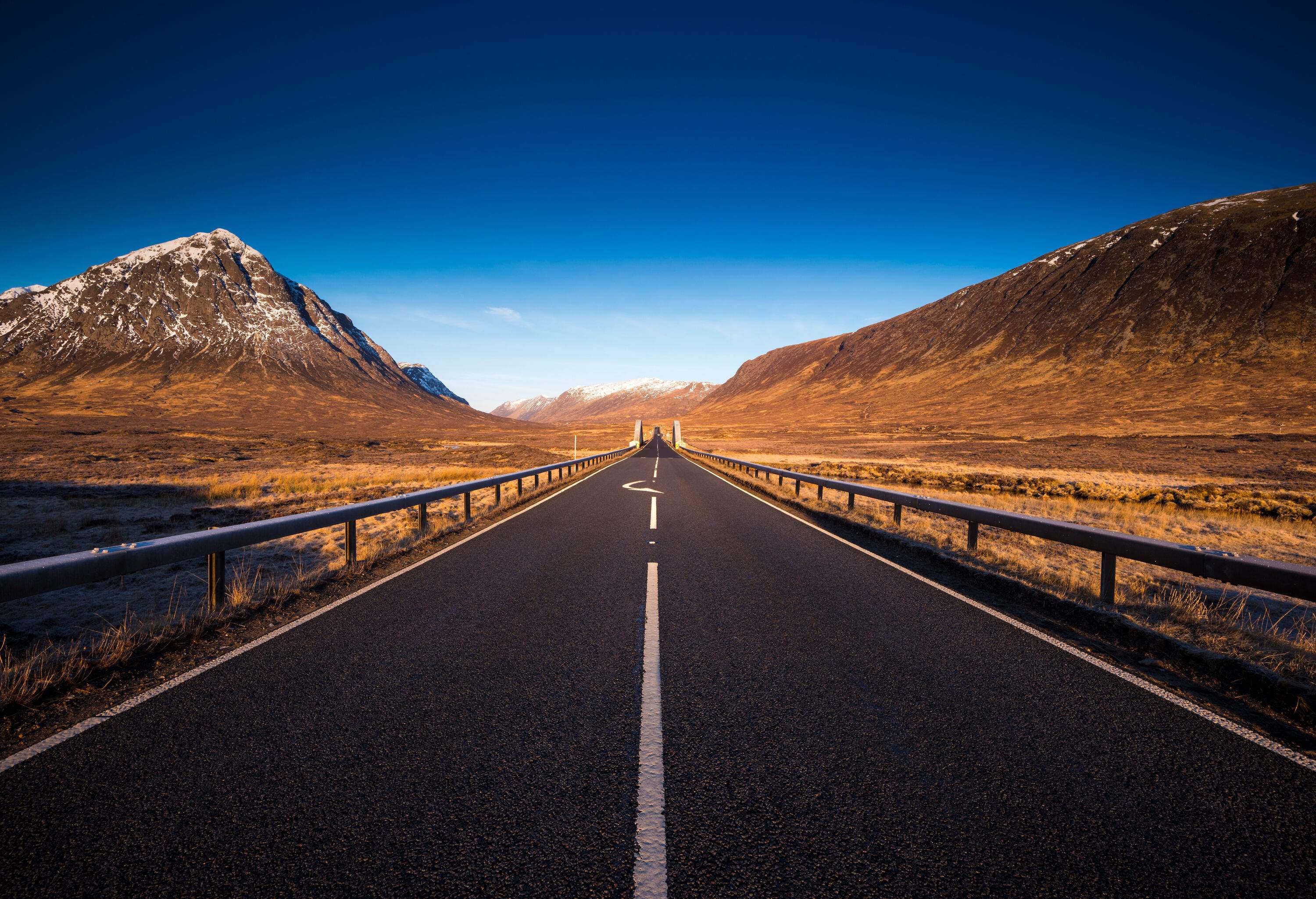 A two-lane asphalt road along a cliff on one side and a cone-shaped mountain on the other towards snow-covered ranges in the distance.