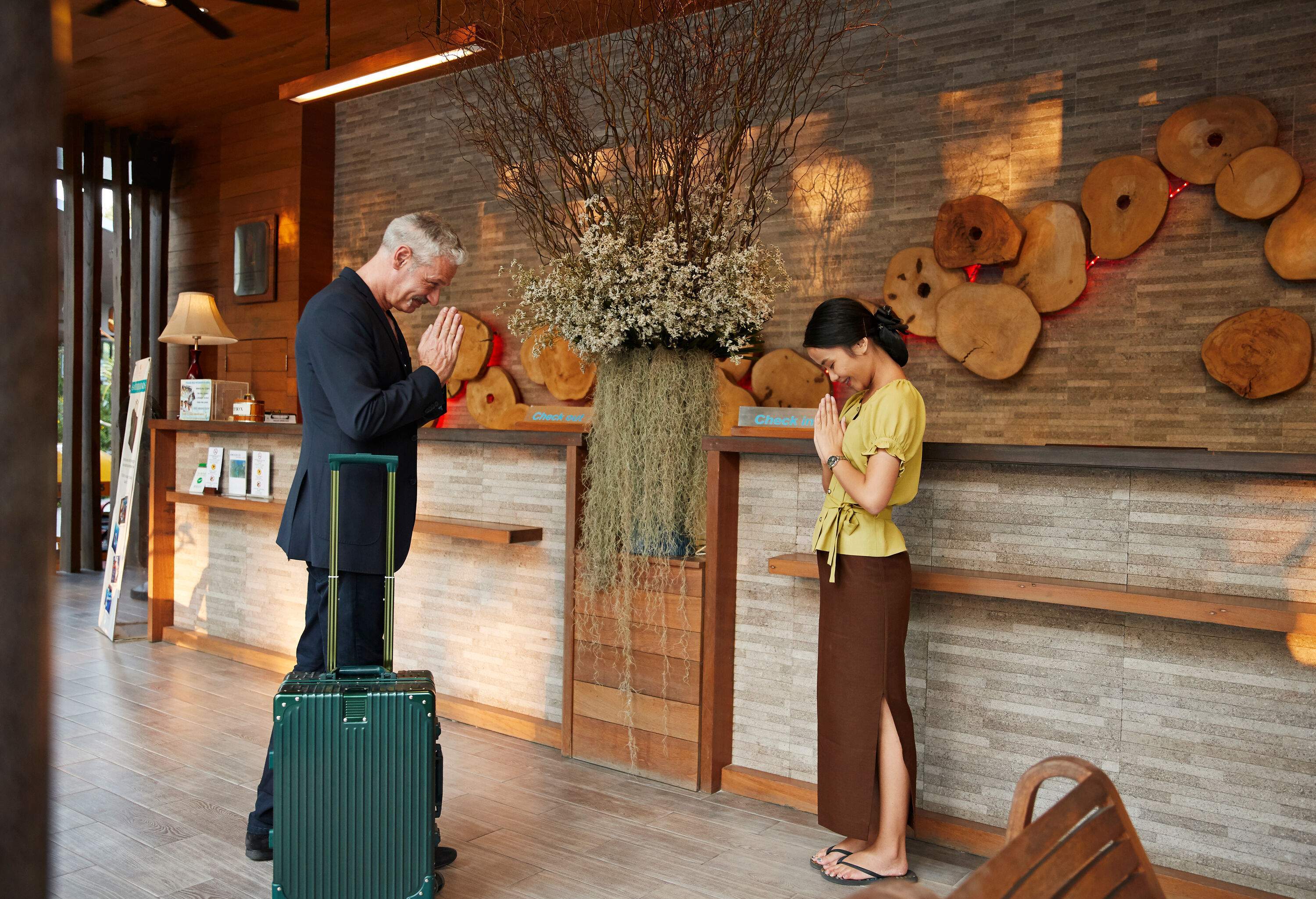 A young female receptionist greeting a male visitor in a traditional wai gesture.