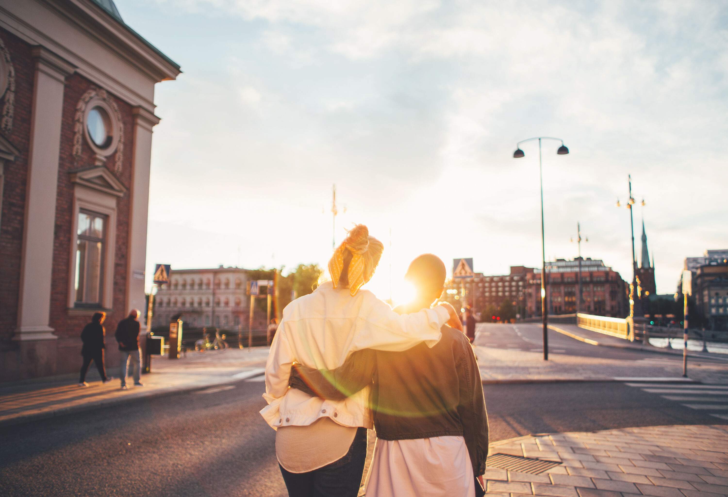 A couple embrace each other at the waist and shoulder as they stare at a sunset.