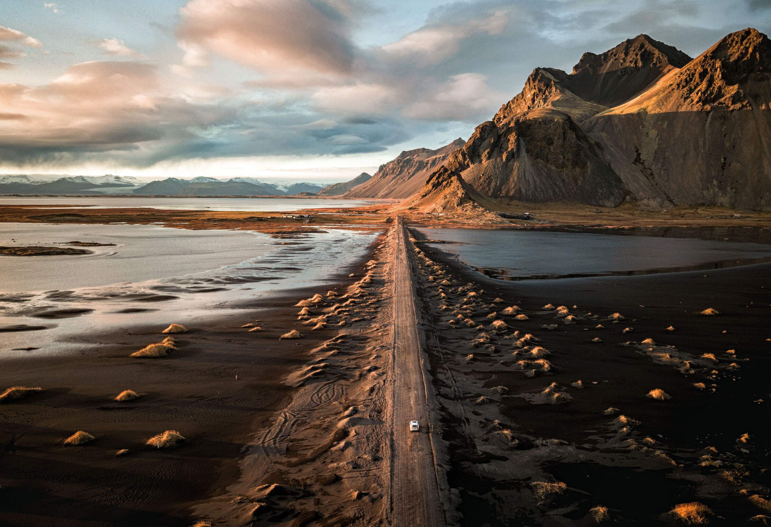 A white car on a rugged narrow road in the middle of black sands heading towards steep pointed mountains.