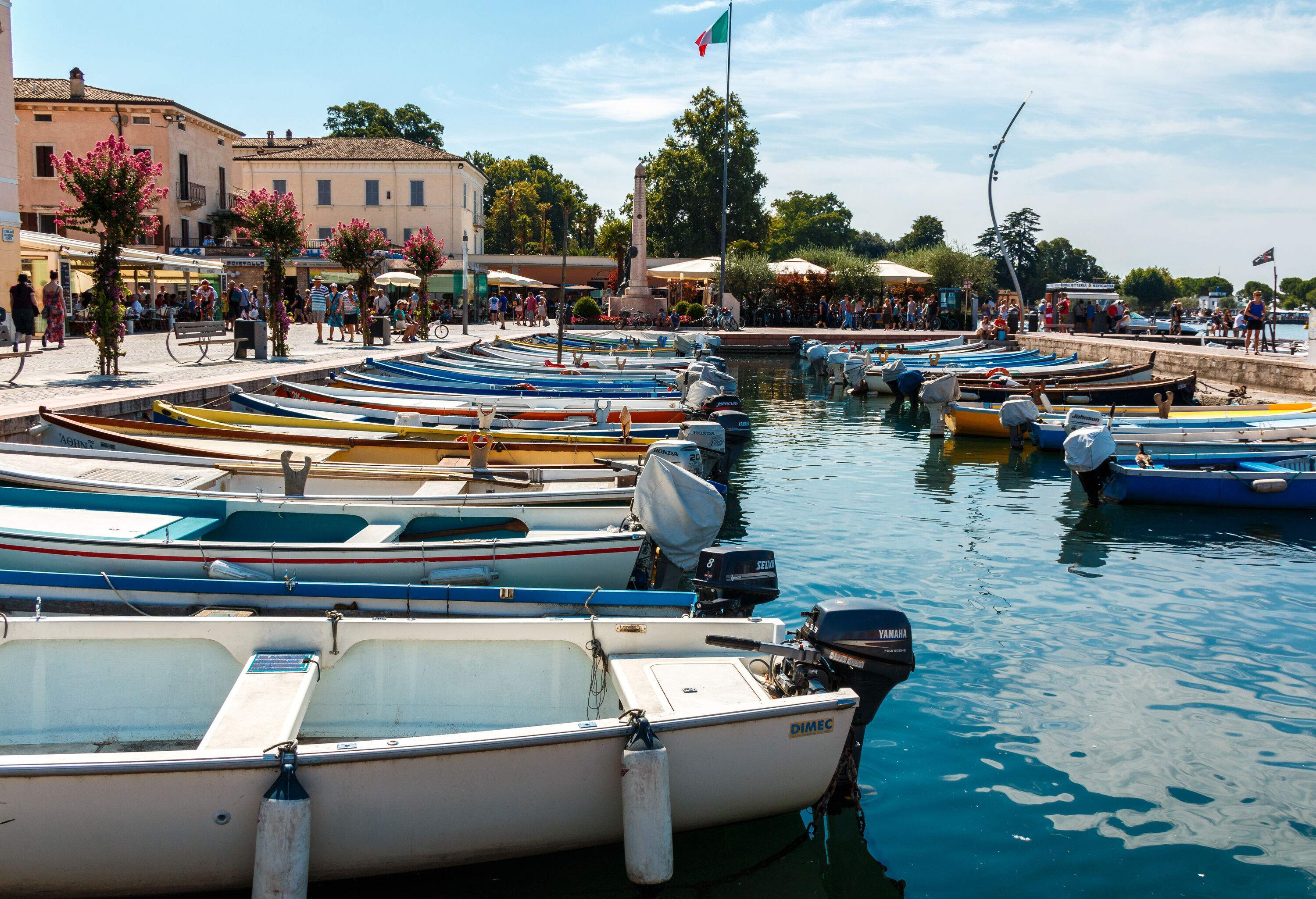 People walk on a harbour lined with anchored boats.