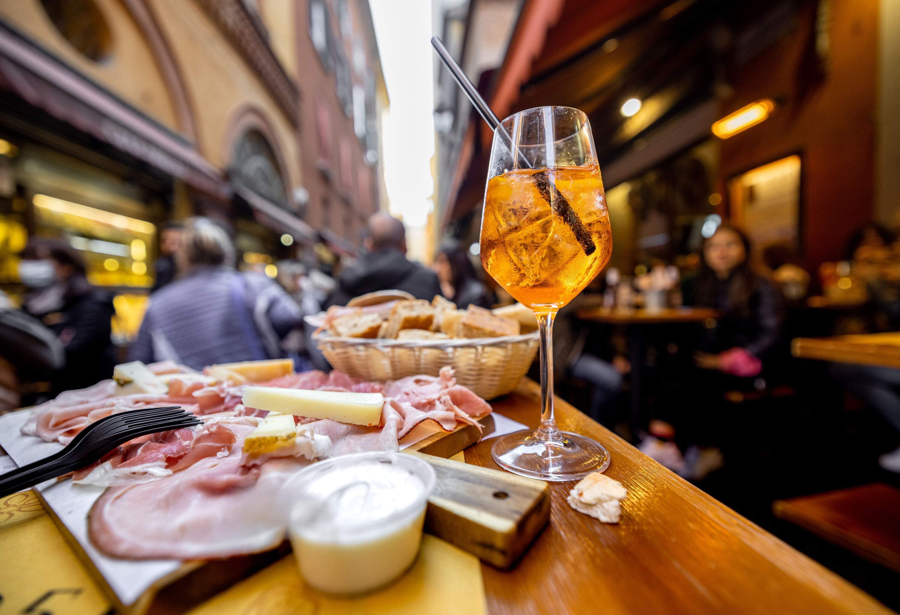 Cured ham in skinny slices, cheese, a basket of bread, and a glass of drink served on the table outdoors.