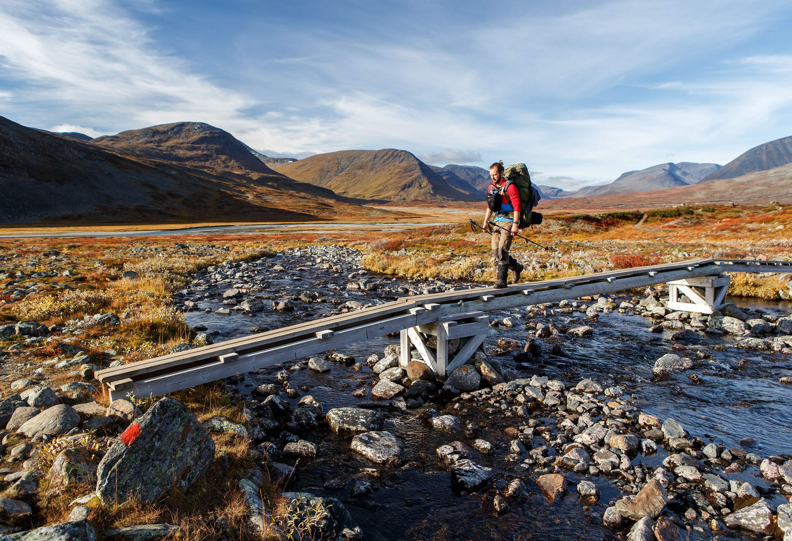 A hiker with his hair tied up crosses a narrow boardwalk on a rocky stream surrounded by tall mountains.