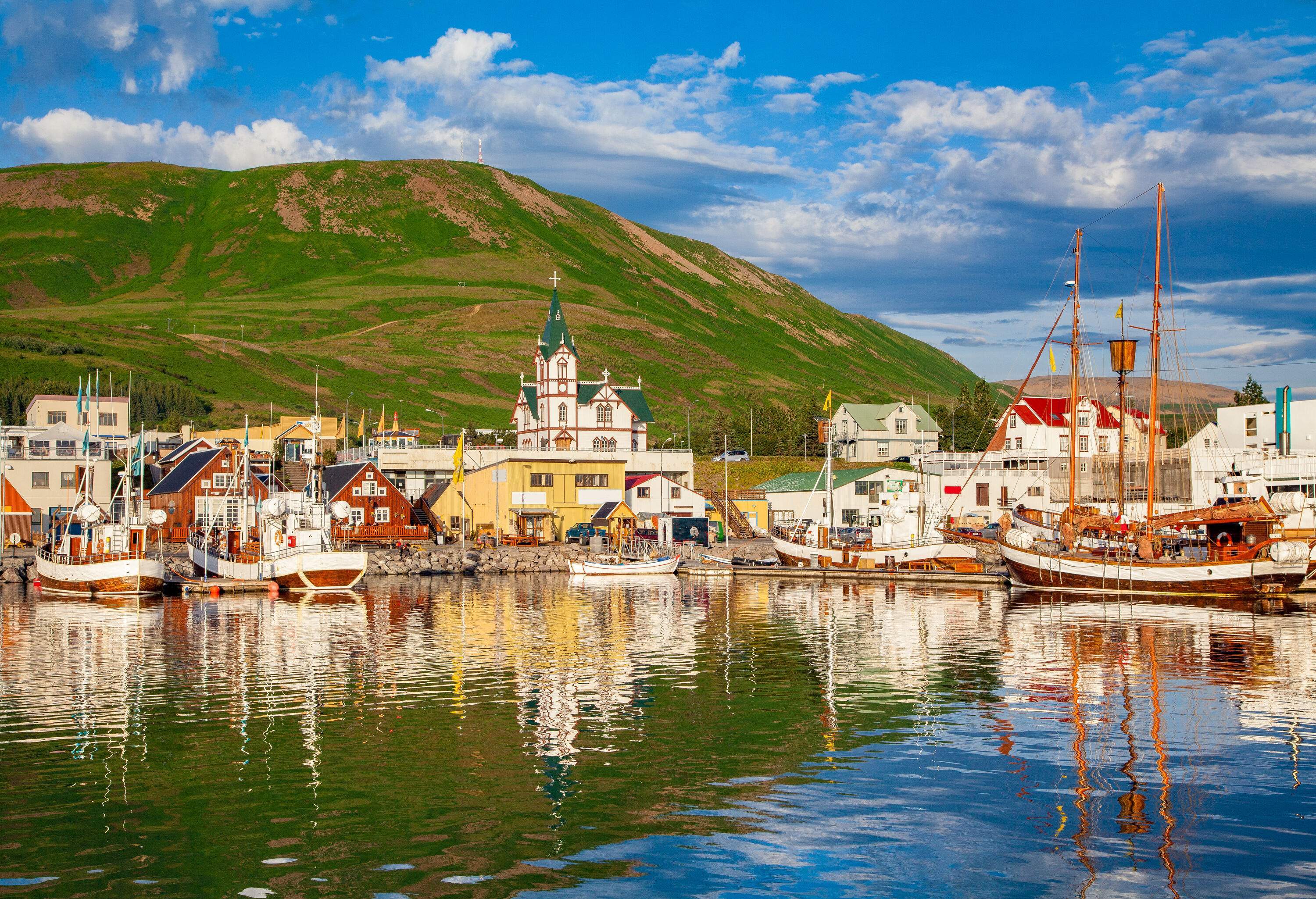 A church sticks out in the middle of traditional houses at the base of a lush green mountain alongside a marina with anchored boats.