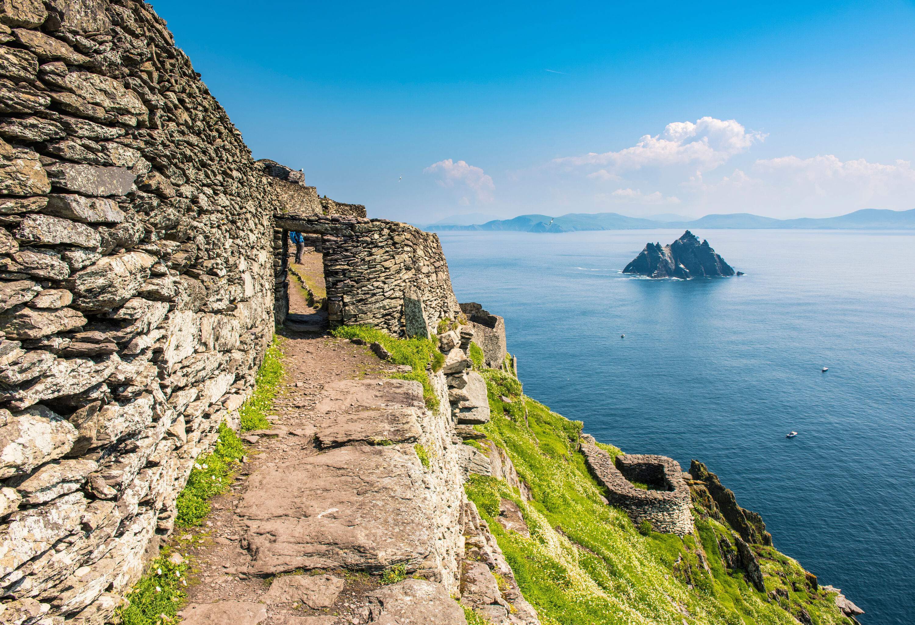 An ancient stone edifice clings to a steep slope, commanding a view of the sea, while a stunning rock formation juts out in the water.