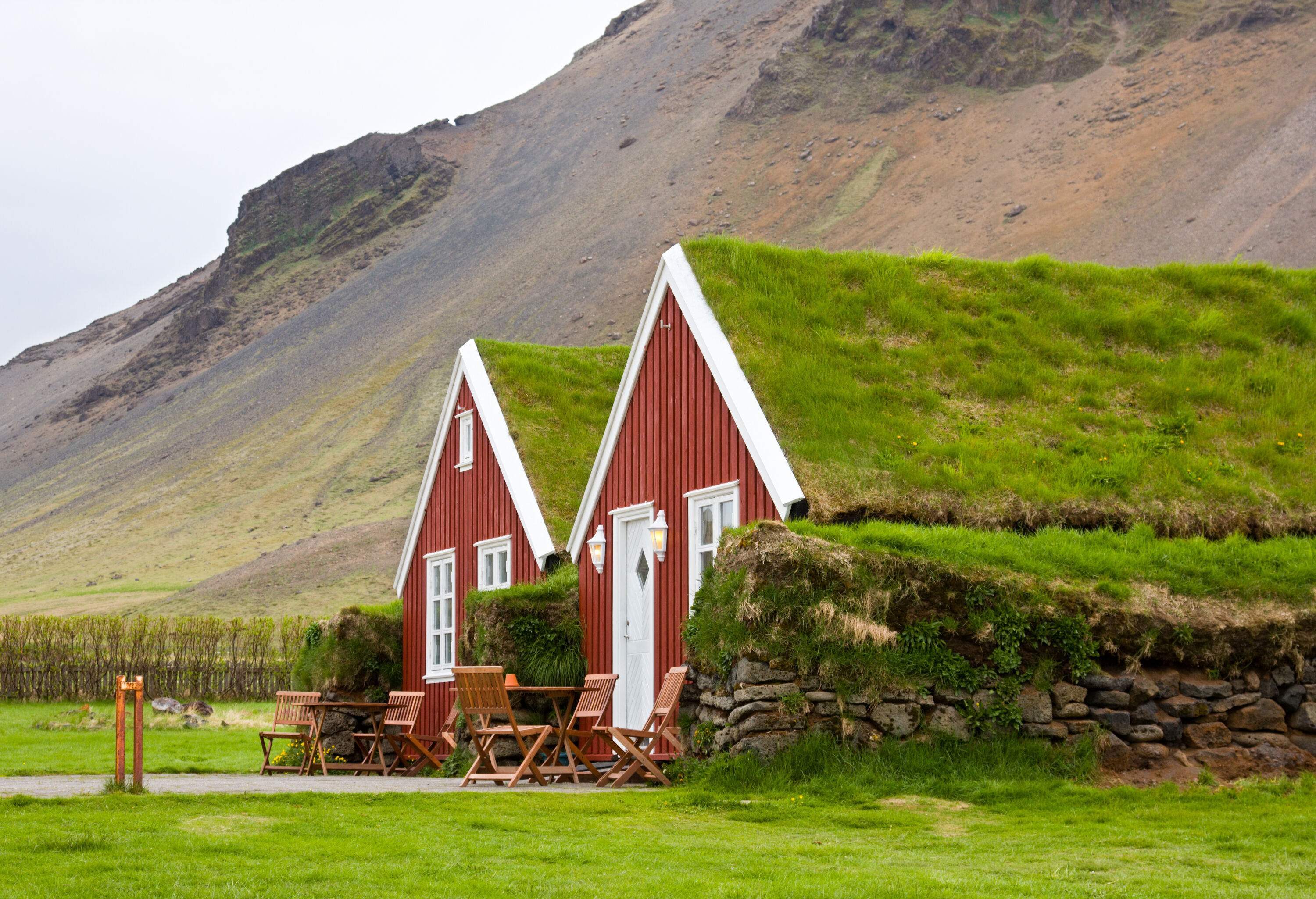 Two lovely red houses with grass-covered roofs are set against a breathtaking backdrop of steep mountains, with wood tables and chairs in front.