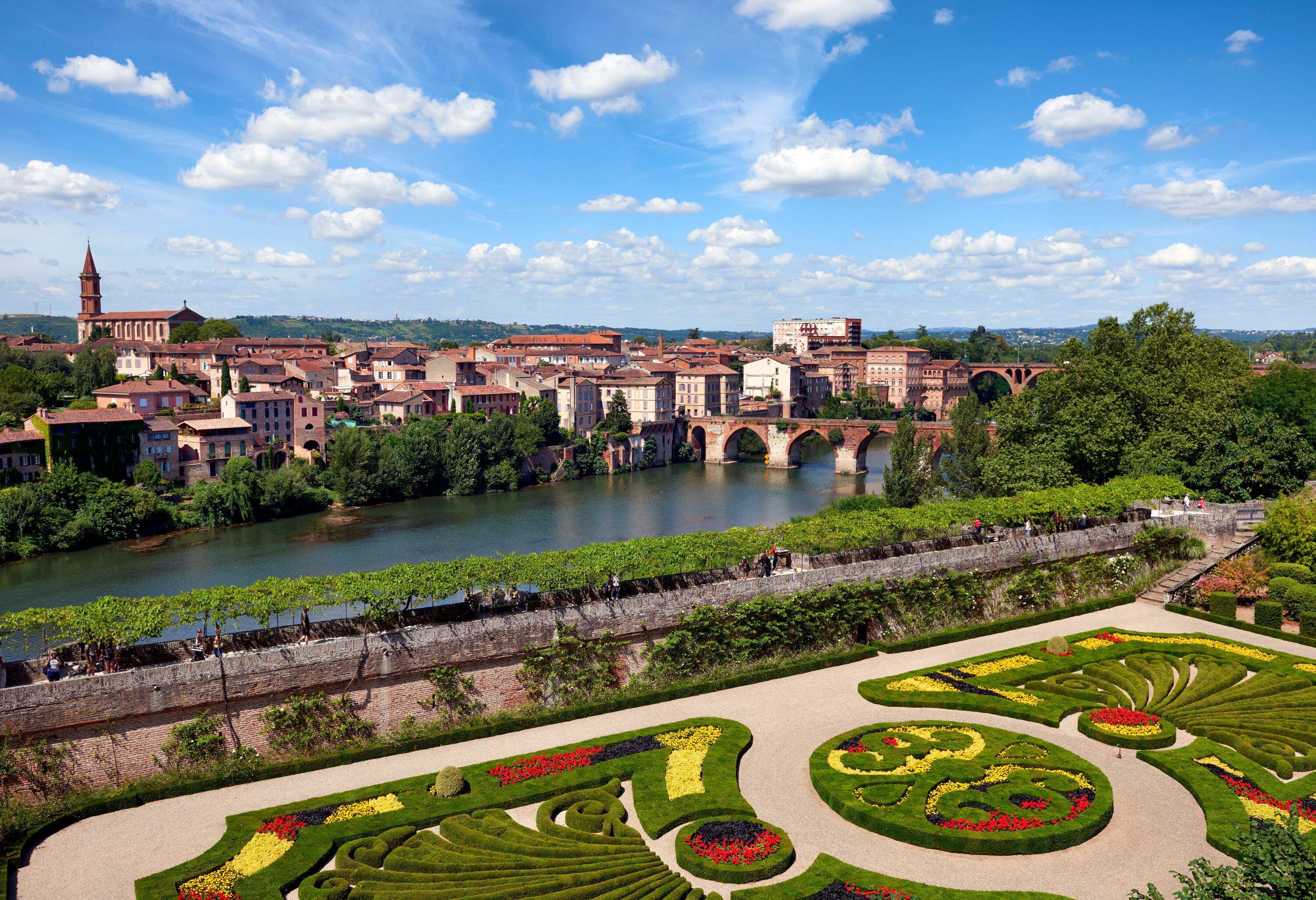 An ornamental hedge garden with an old town and a river bridge in the backdrop.