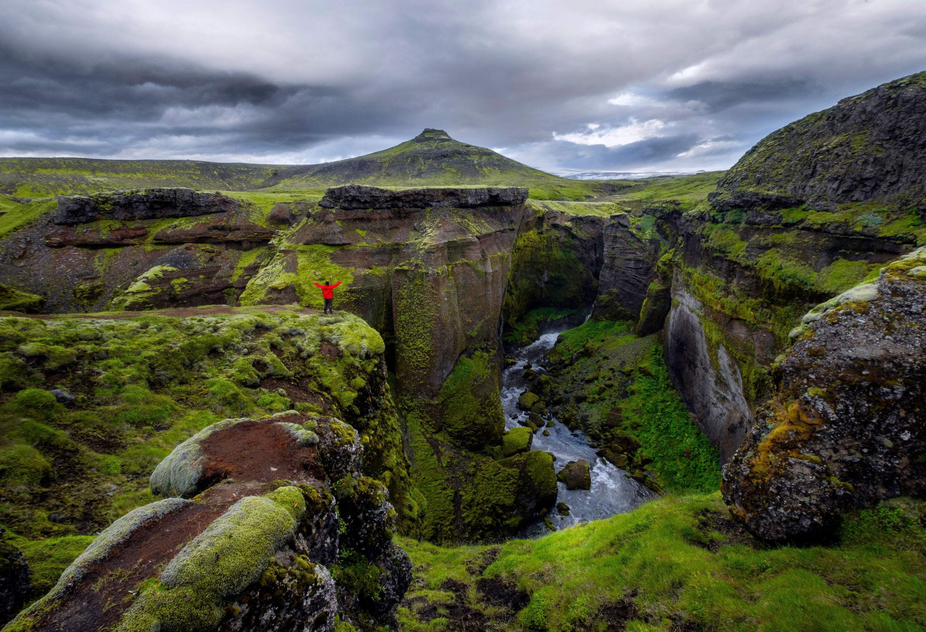 A man in a red jacket with raised hands stands at the edge of a lush, rugged gorge and a stream running through it with a view of a scenic volcano.