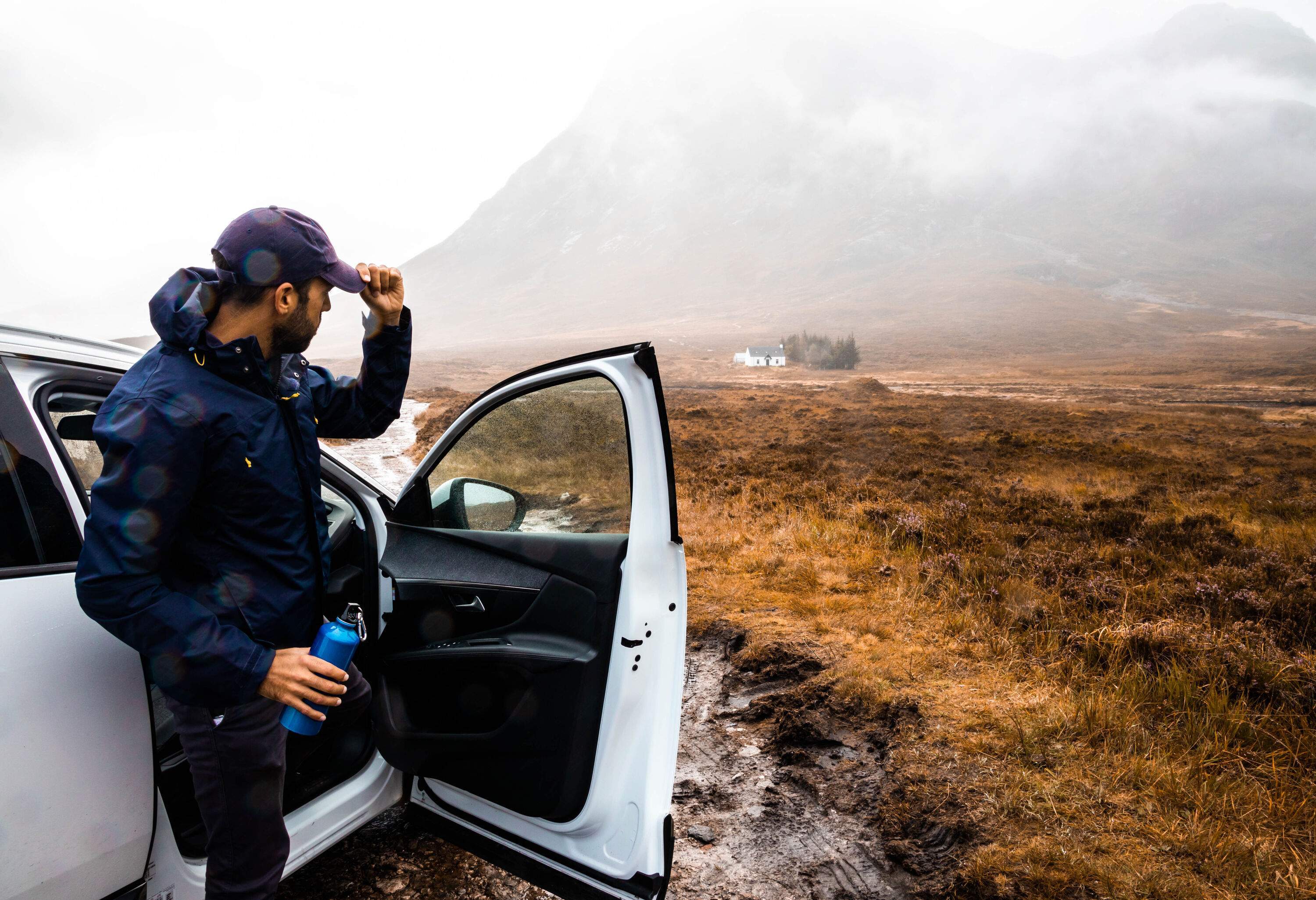 Man clad in a blue windbreaker emerges from a white car, holding a flask and looking at the bare landscape on a cloudy day