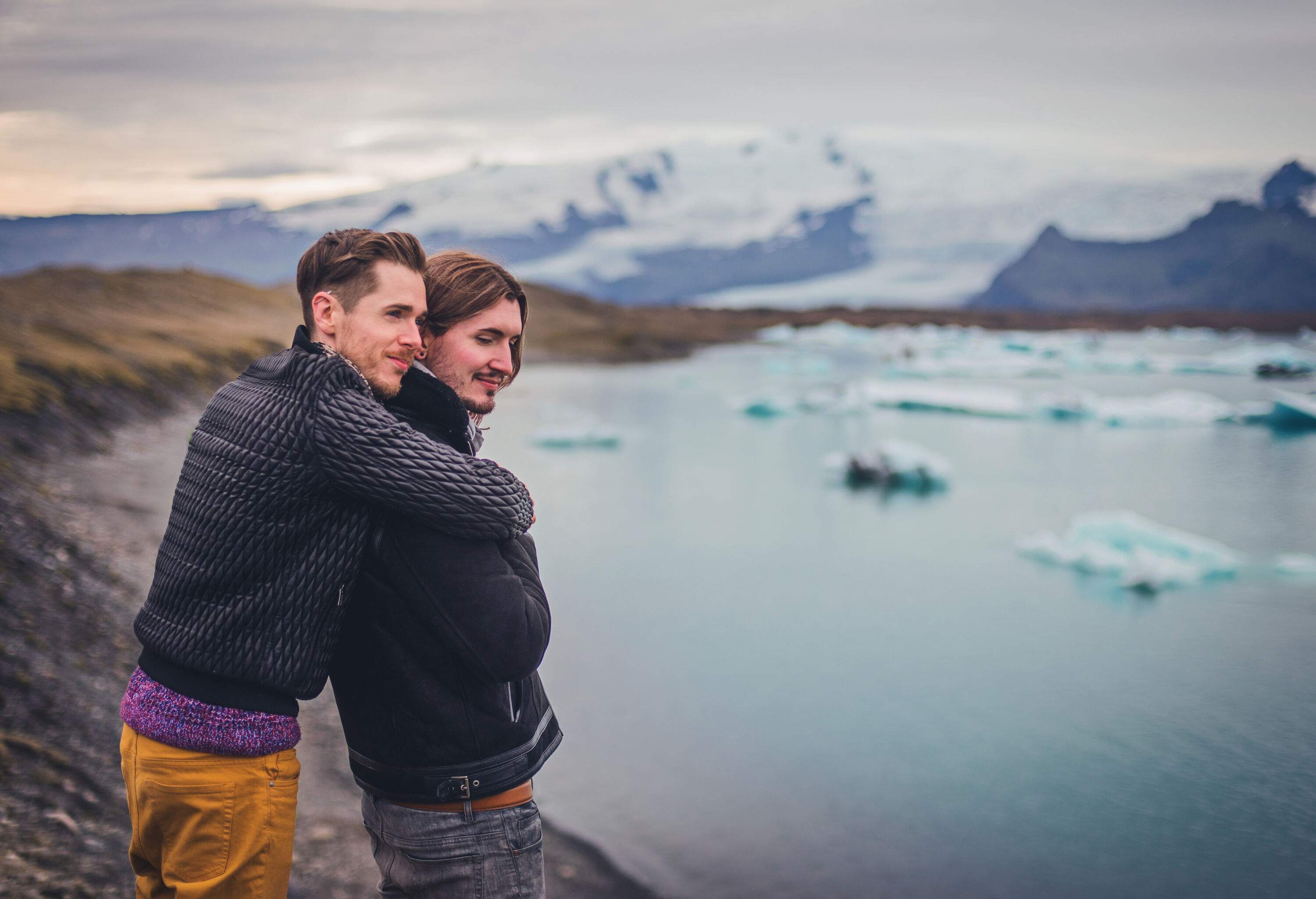 Two smiling men standing on the shore of a lake and hugging with a view of a snowcapped mountain in the background.