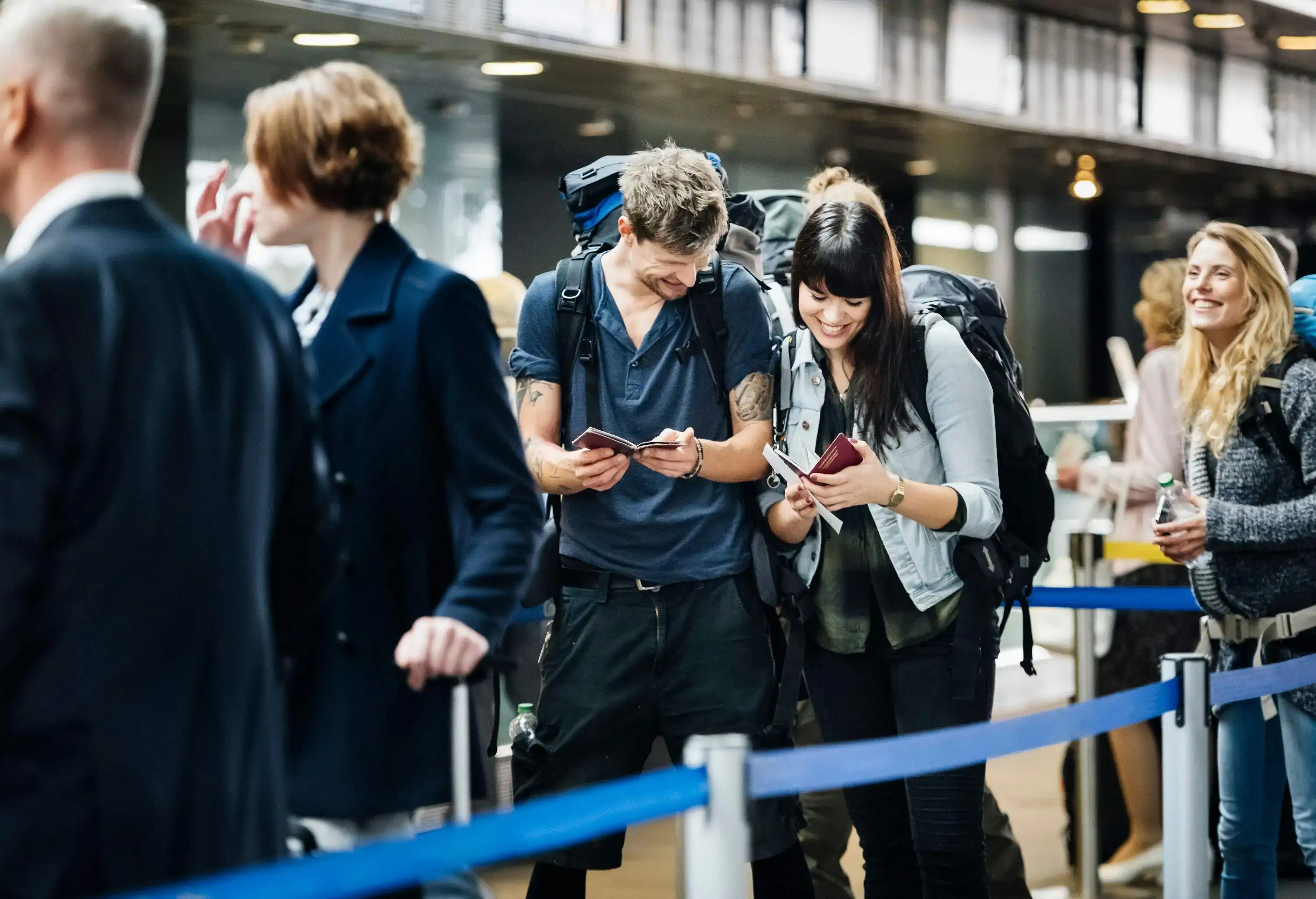 A smiling young backpacking couple eagerly checks their passports while waiting in a queue.