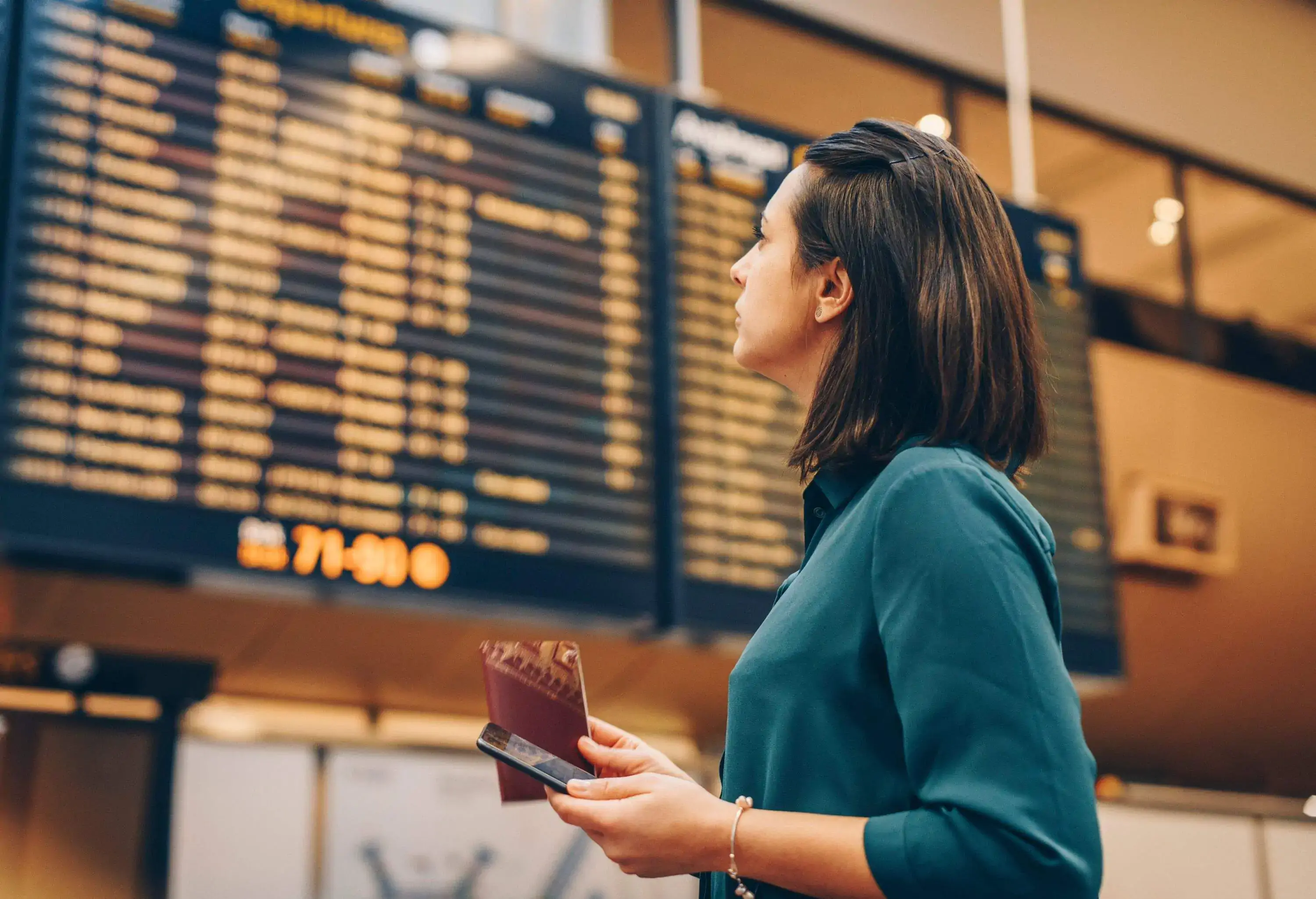 portrait of woman wearing green shirt looking at airport flight board holding a mobile device and passport