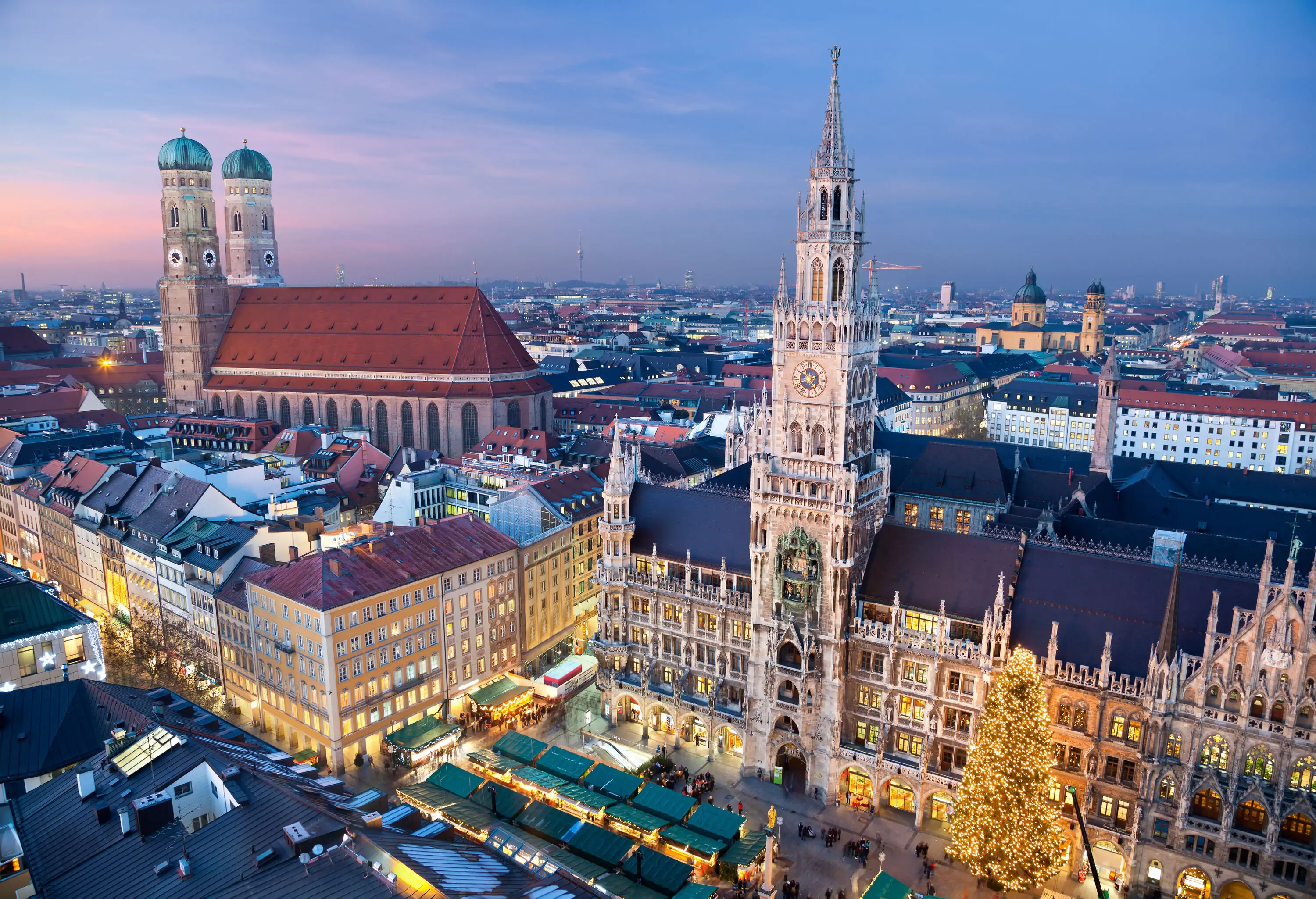 Christmas trees and shops on a public area along the Gothic Revival-style New Town Hall, with views of the Frauenkirche, a red tiled church with domed spires.