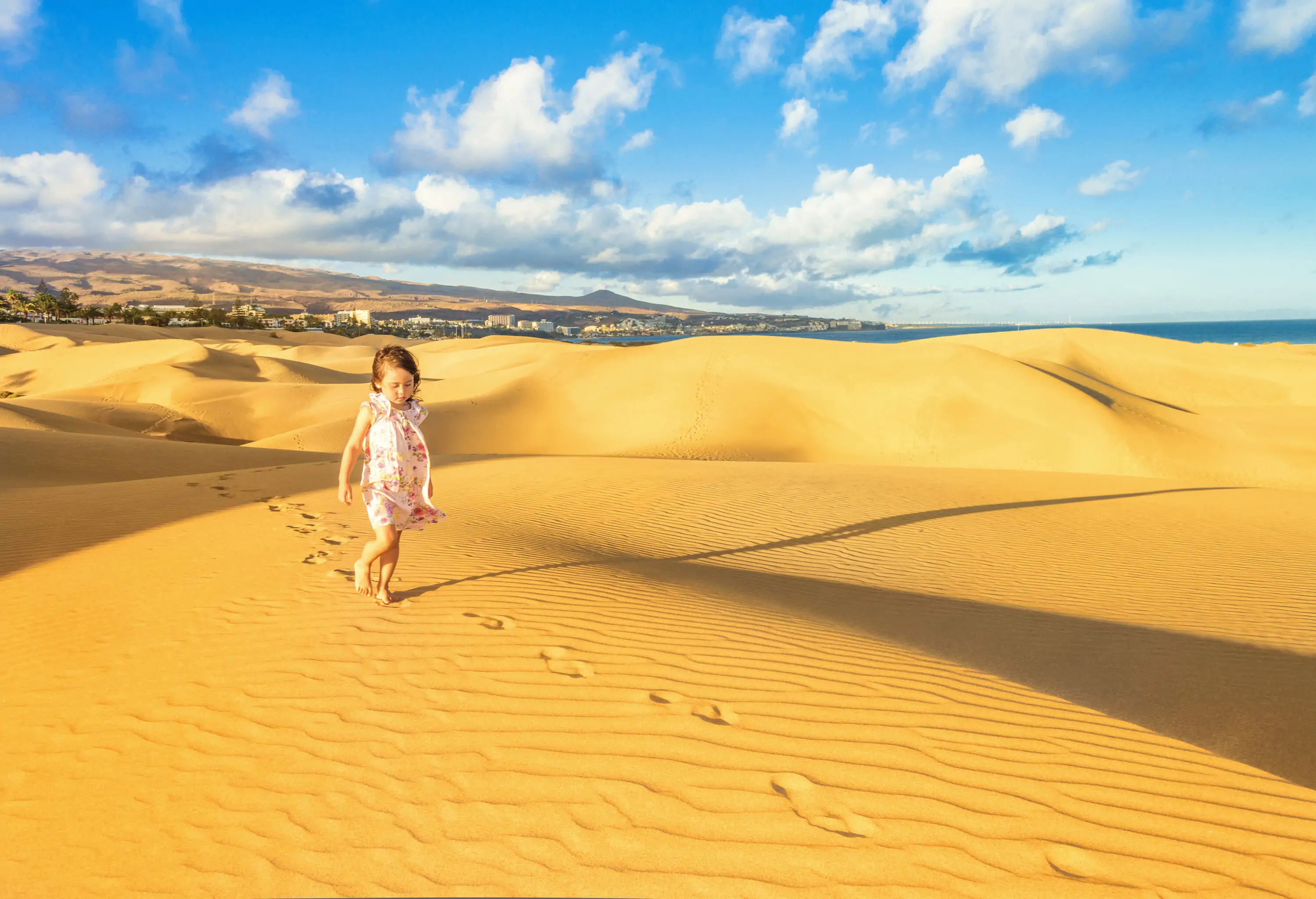 A little girl in a pink floral dress following a trail of footprints on sand dunes.