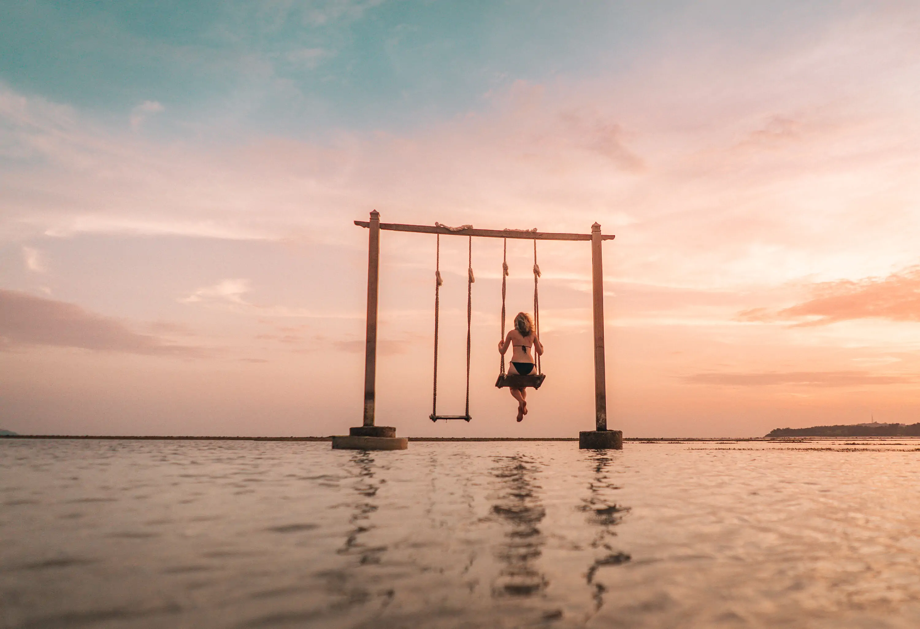 A lady in a black bikini is sitting on the swing in the sea's shallow waters on a scenic sunset.