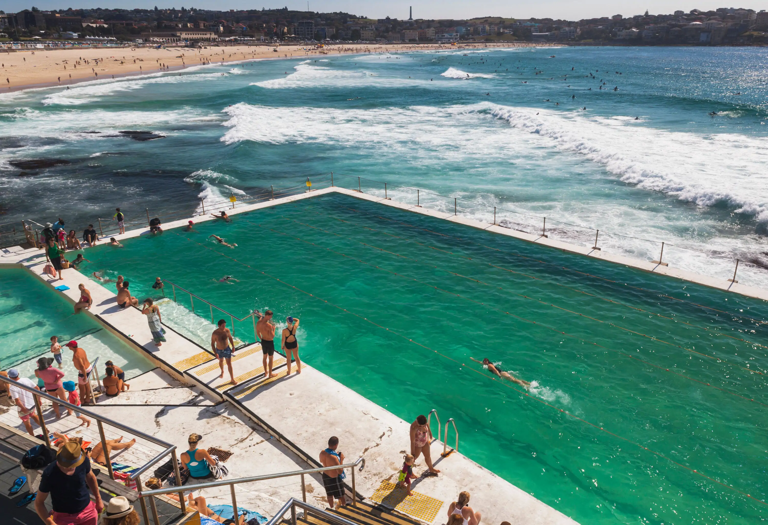 Many people are on the beach enclosed by mountains and in the pool next to the sea with the rolling white waves.