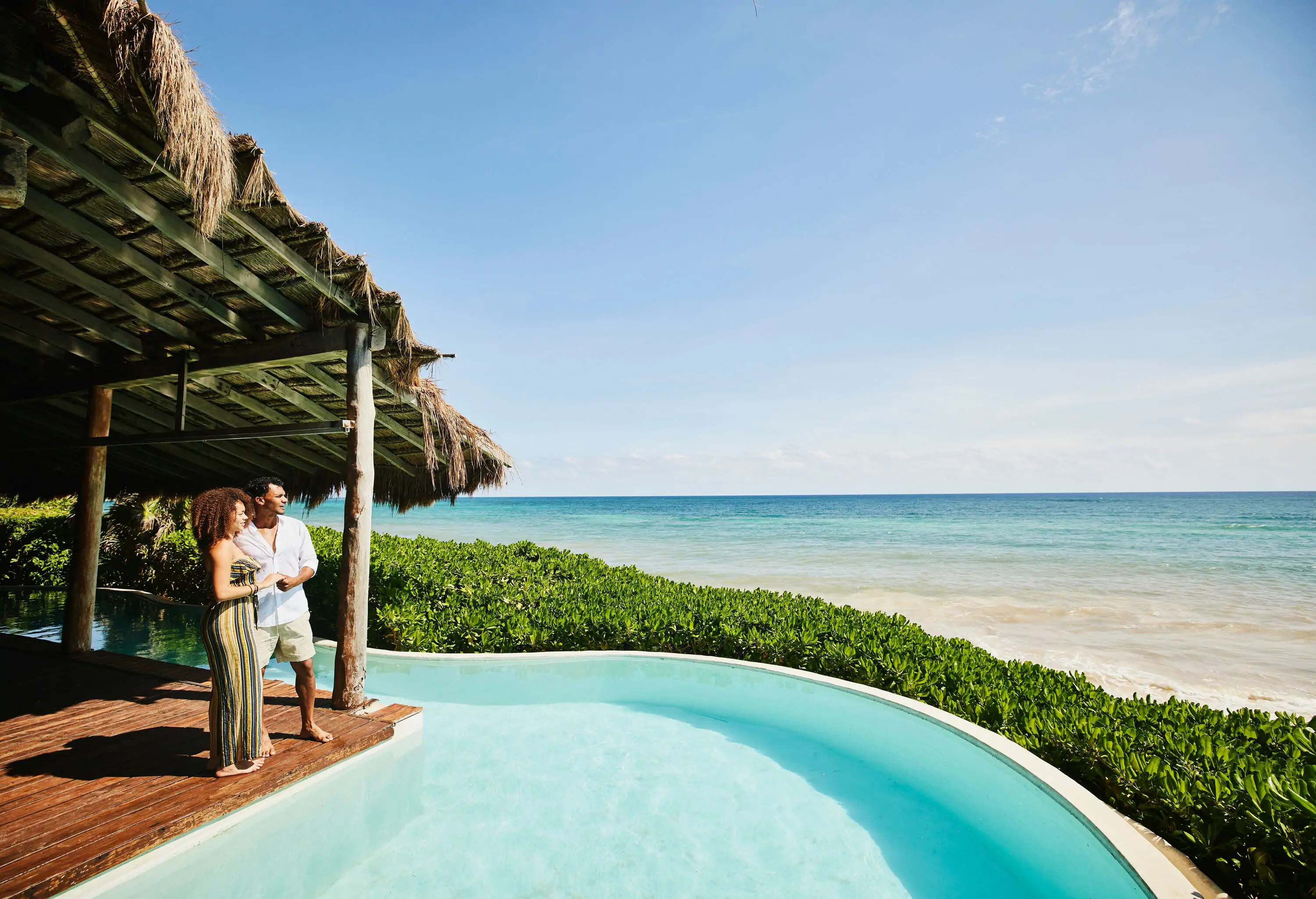 A couple finds solace together, standing side by side under a thatched shade at the edge of a pool.