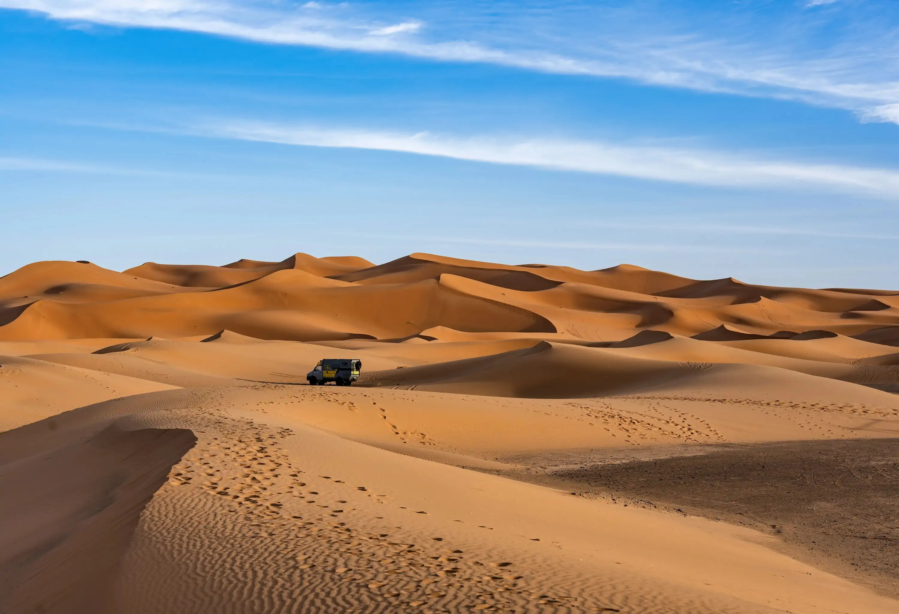 An off-road vehicle navigates the sweeping dunes of the Sahara under a wide blue sky, showcasing the allure of adventure travel.