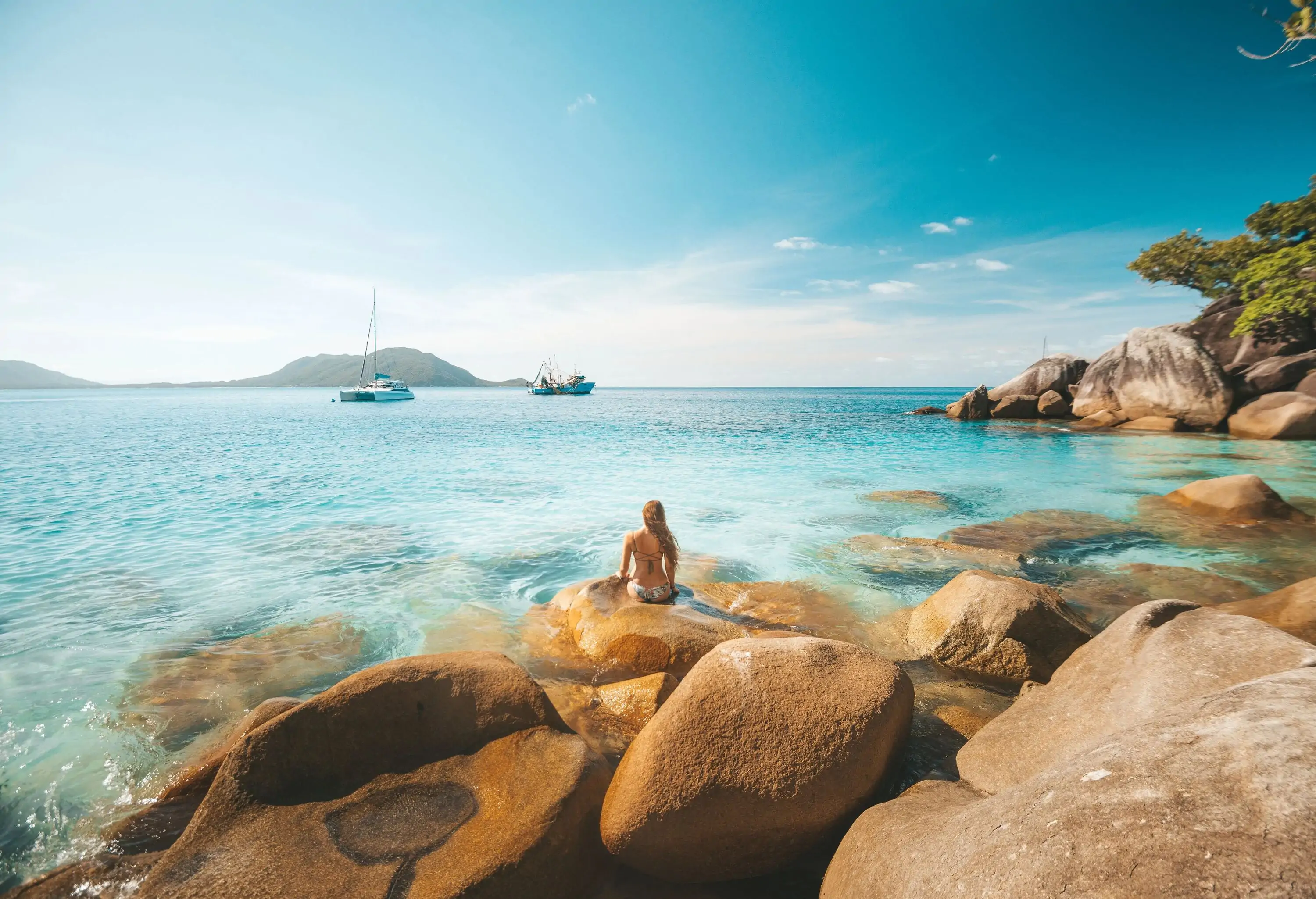 A woman in a bikini sits on coastal rocks admiring a vast ocean with cruising boats.