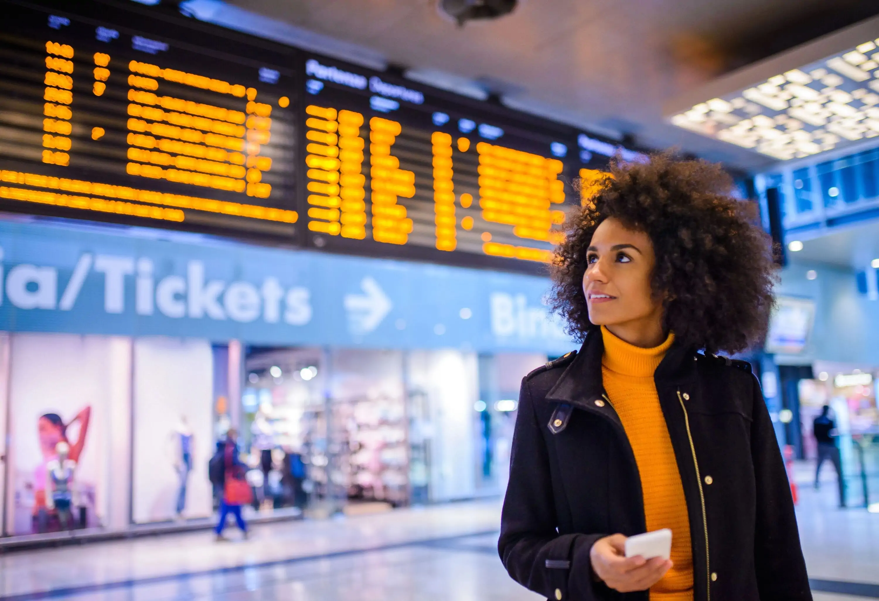 A woman with beautiful afro hair looks up as she holds her mobile phone against the backdrop of a bright airport.