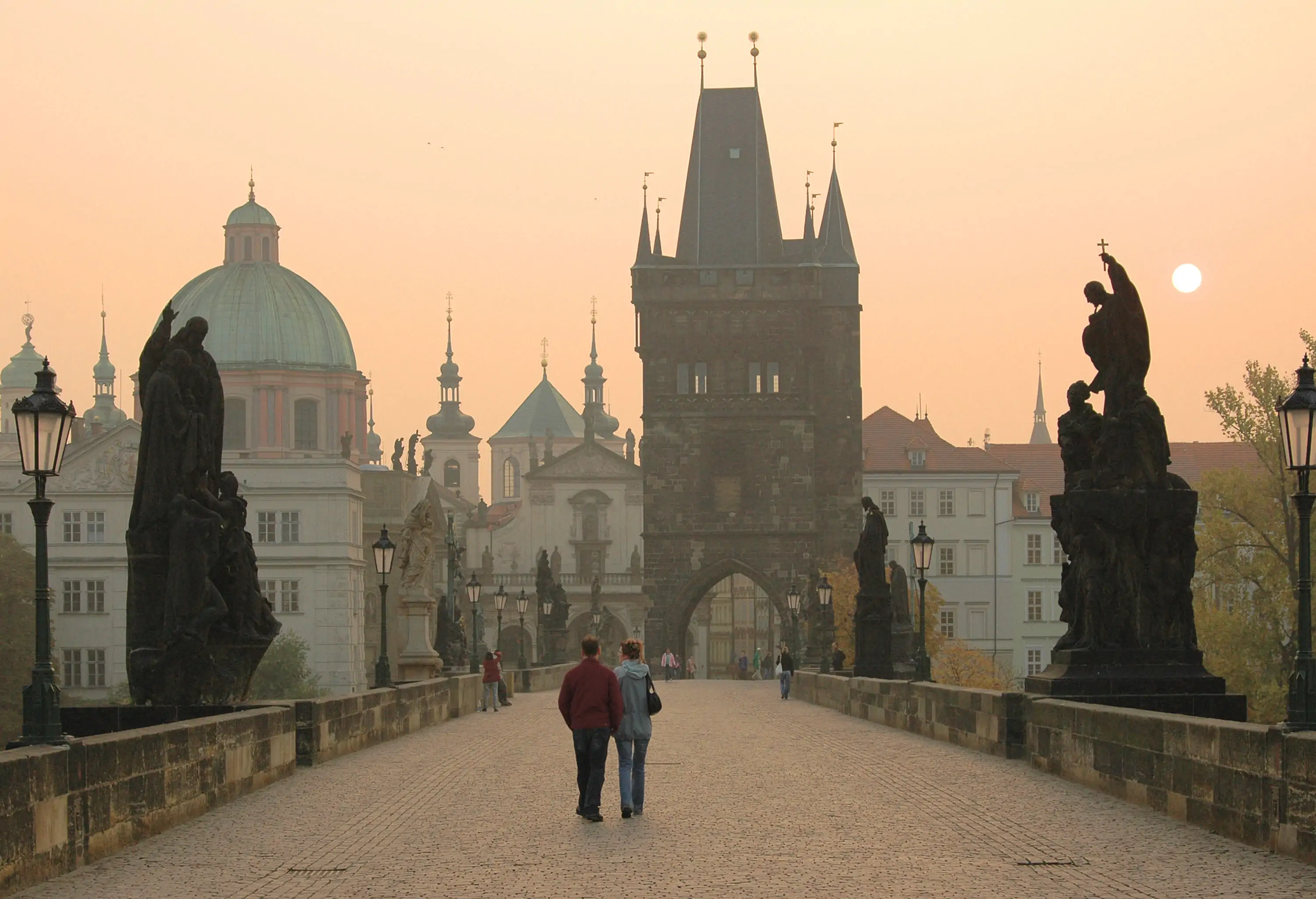 A couple walking so close together across Charles Bridge with under a pinkish sky.