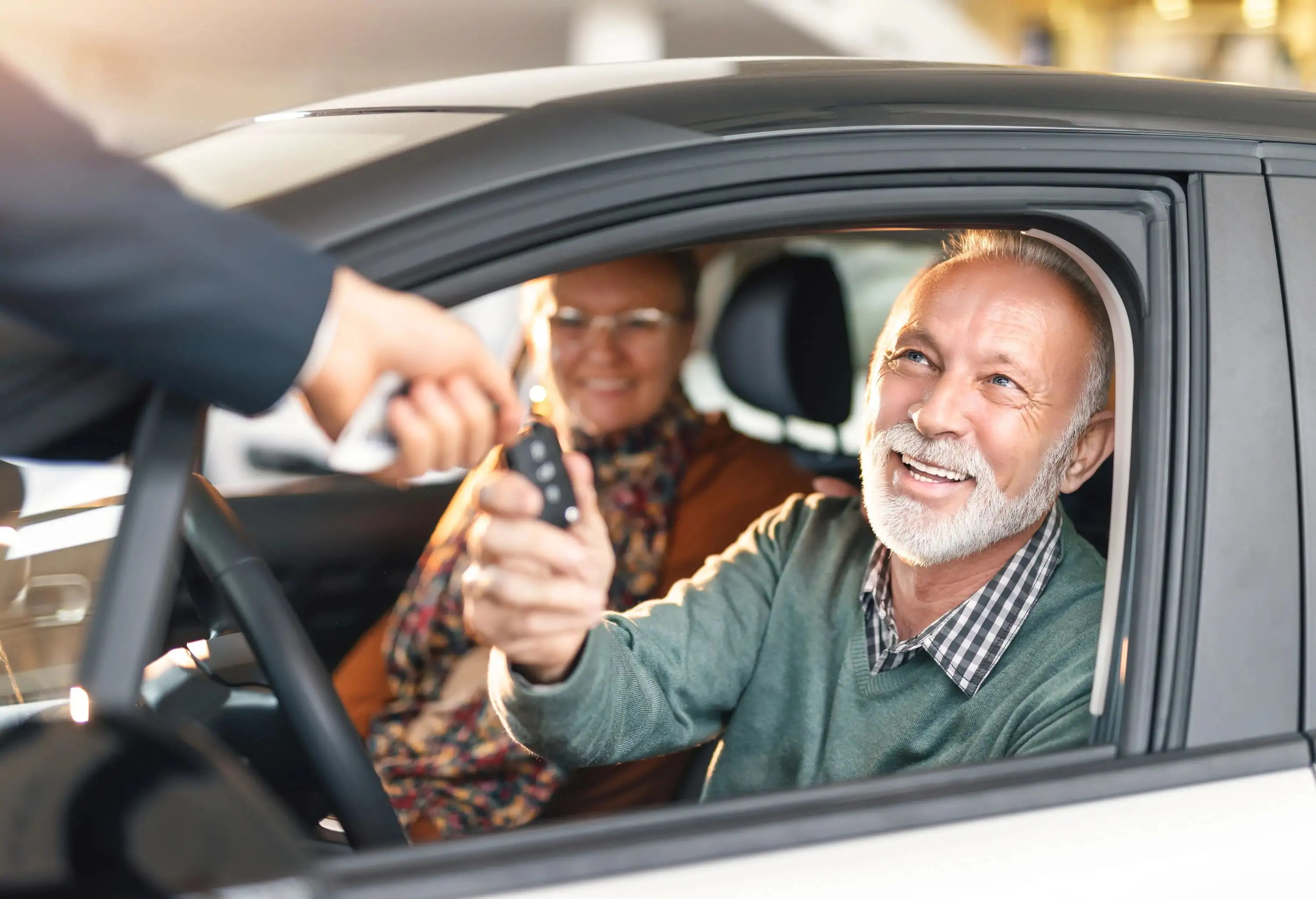 A senior man sitting in the driver's seat reaching for the car keys from someone.