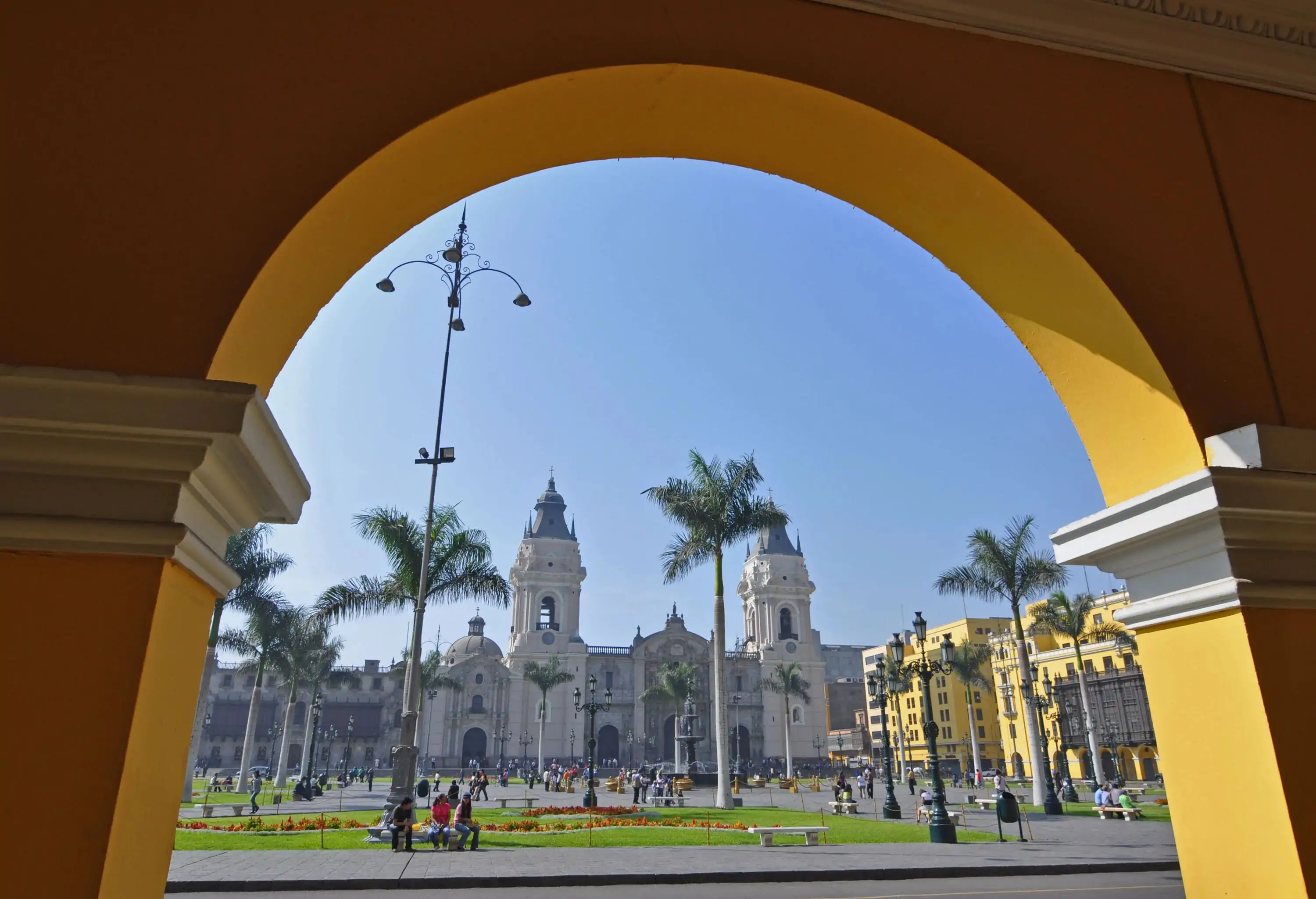 A yellow-painted archway framing a church and a plaza with palm trees and green grass.