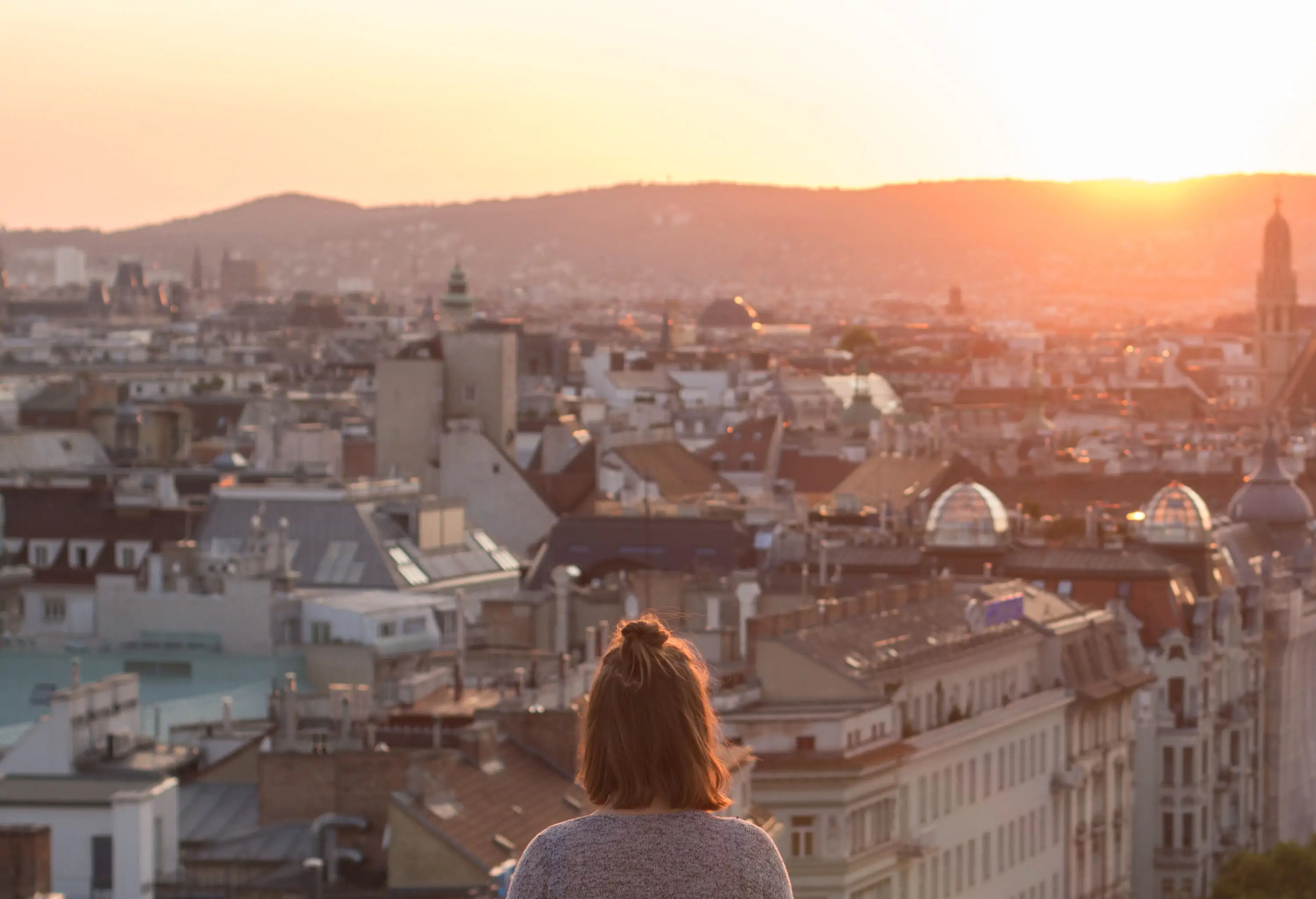 A woman looking out over magnificent city buildings as the sun peeks over the mountains.