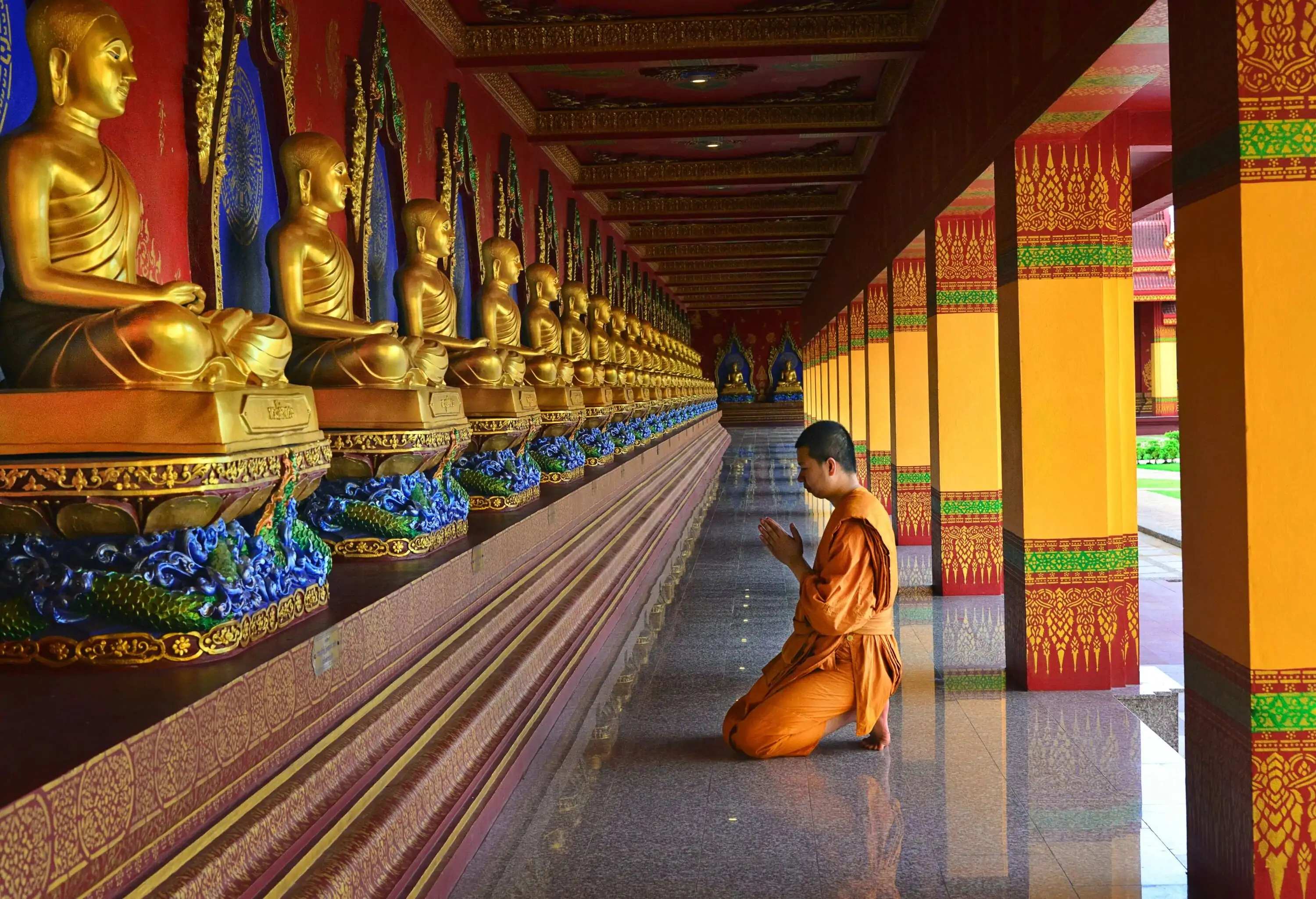 A devout monk kneels in prayer before a row of resplendent golden Buddha statues, surrounded by intricately designed columns that attest to the sacredness and artistry of the spiritual space.