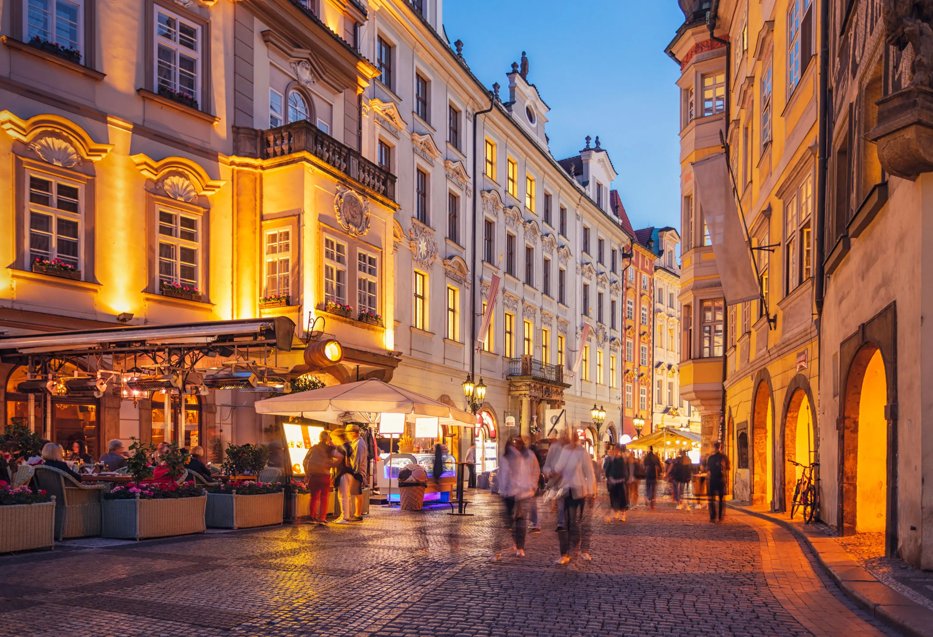 A night-time view of a busy street lined with outdoor dining areas and brightly lit commercial establishments.