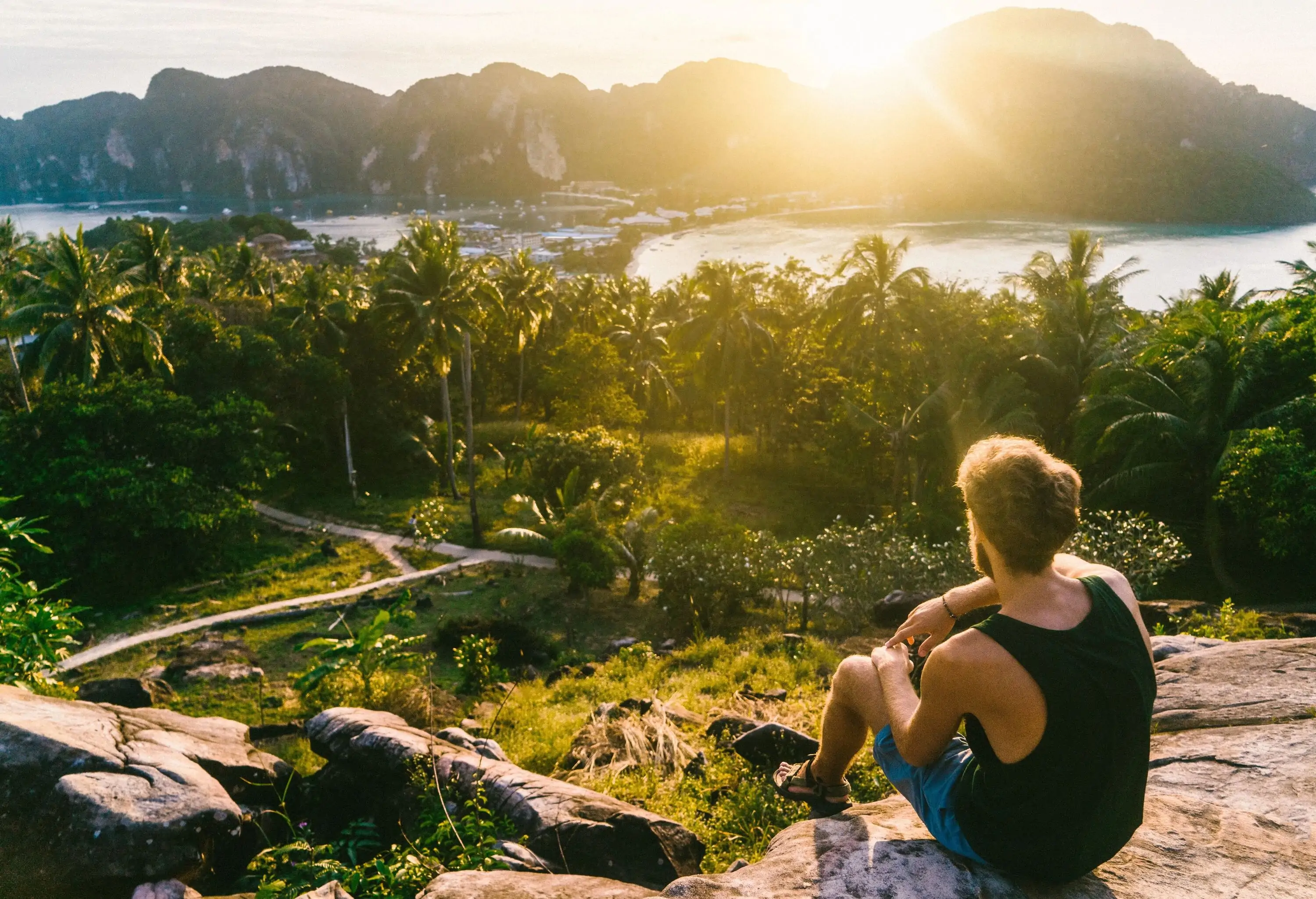 A young Caucasian man gazes out at the breathtaking view of a bay from atop a hill, surrounded by lush tropical trees, immersing himself in the beauty of the natural scenery.