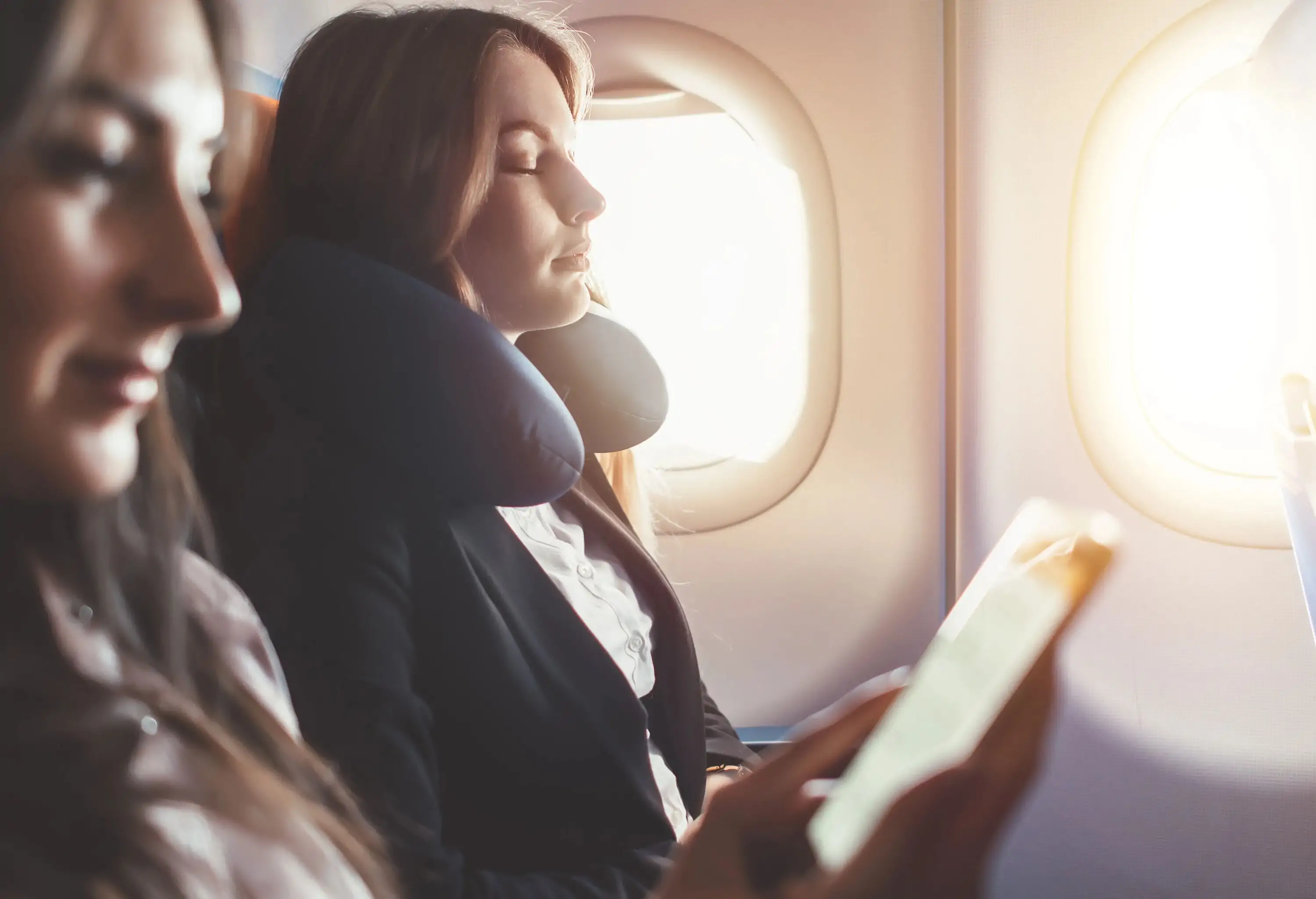 A woman closes her eyes as she takes a nap in an airplane seat beside a woman using a smartphone.