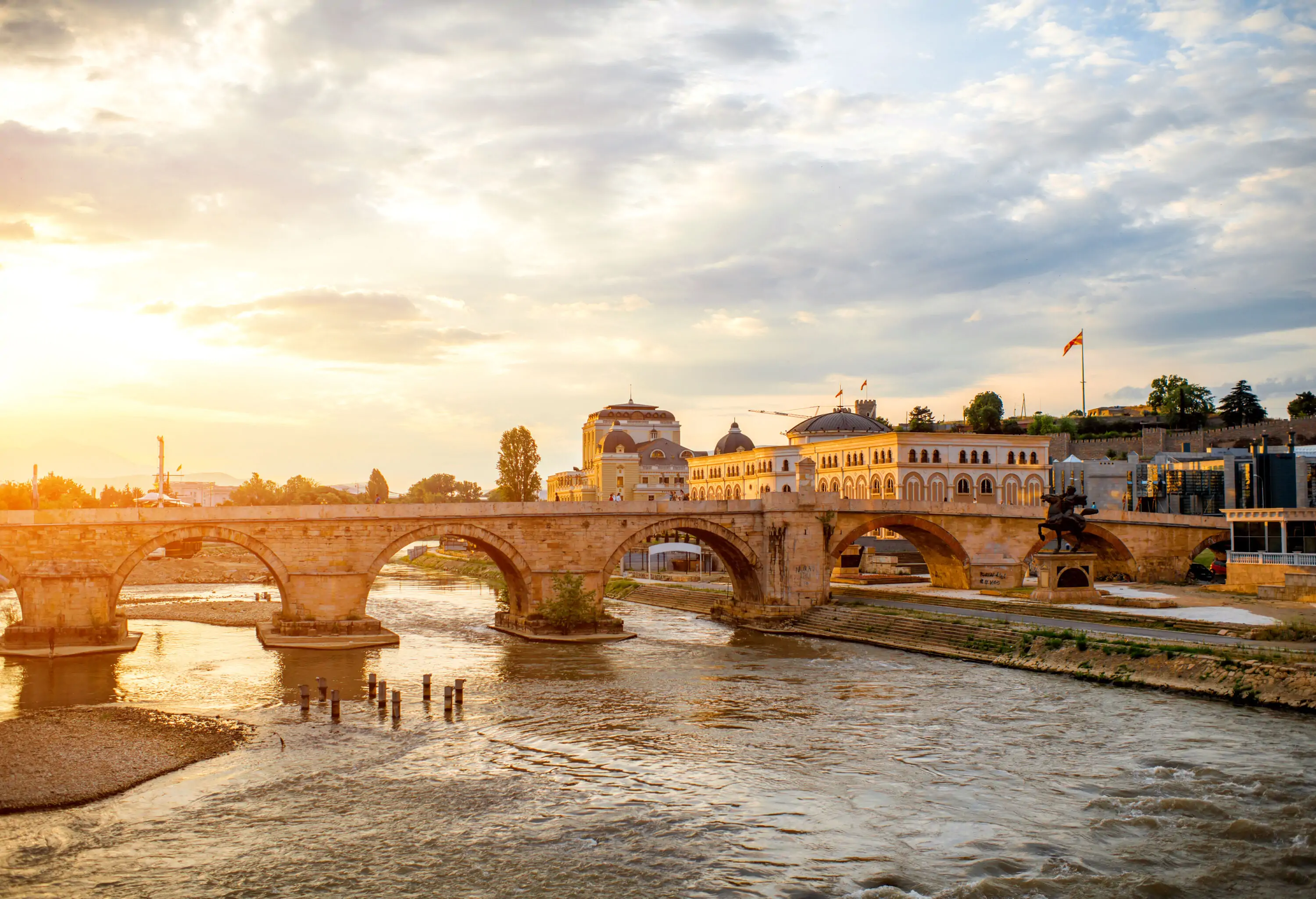 The scenic sun shines through a stone arch bridge that spans the river along the old buildings.