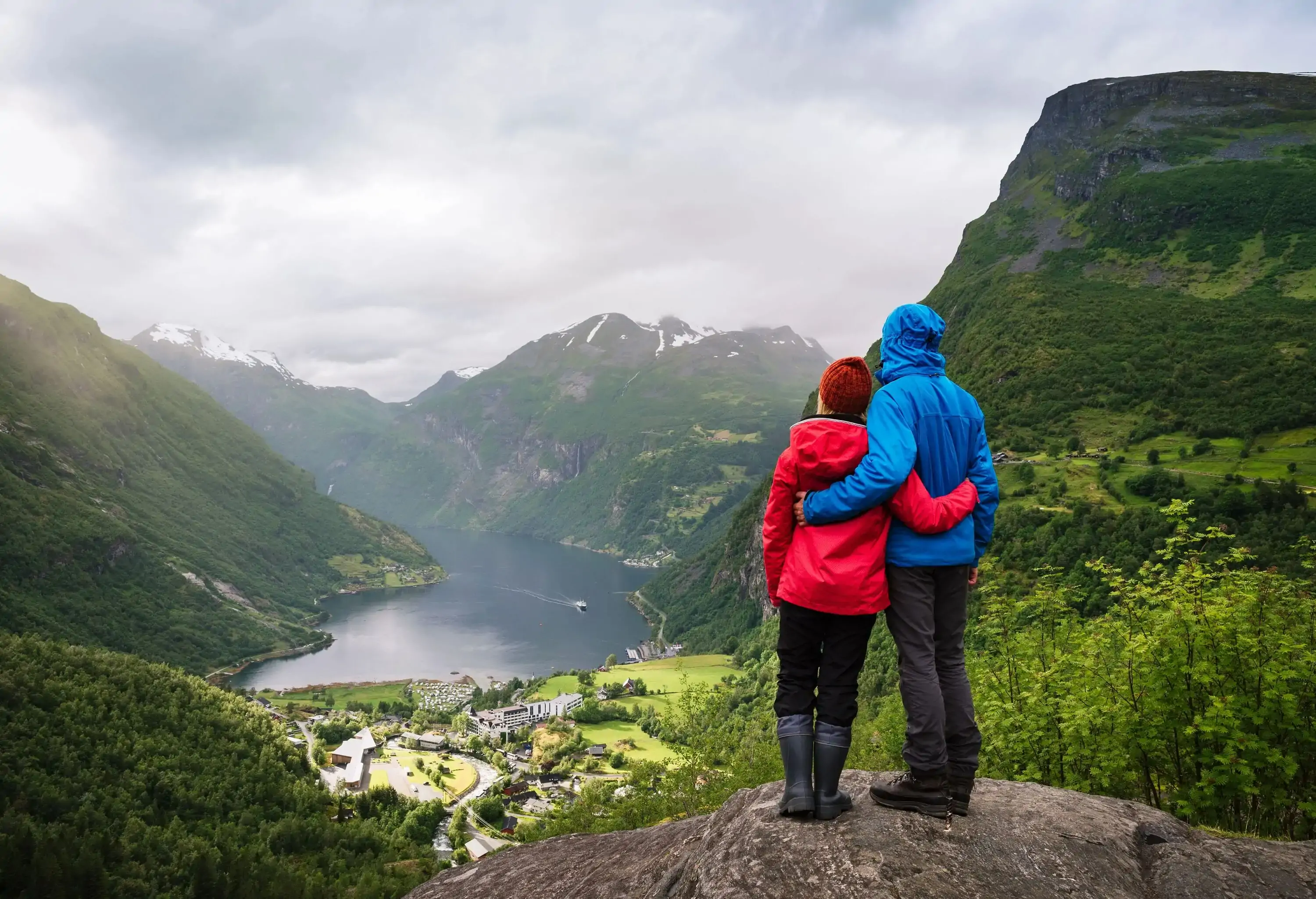 A couple in red and blue jacket stands on a rock and looks at a village along a lake and forested slopes.