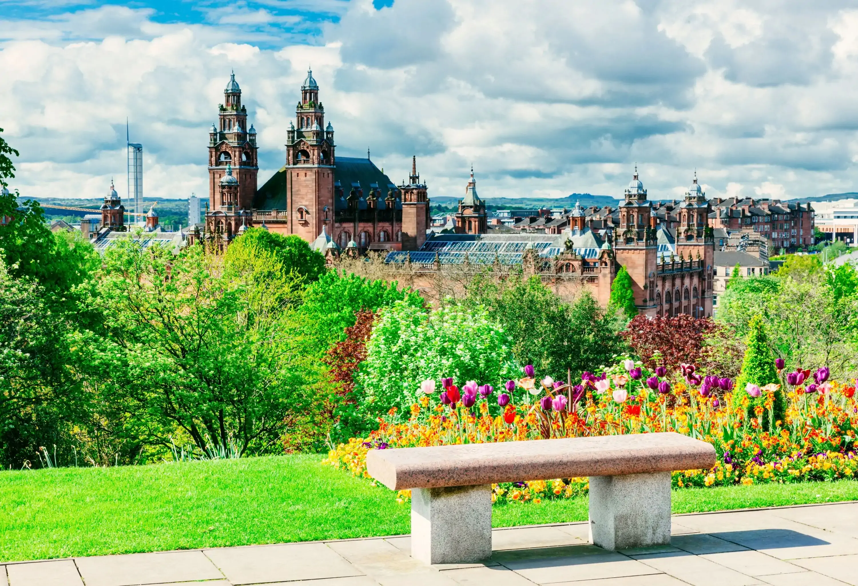 A Gothic Revival architecture University building is seen from a lush park with colourful flowering plants.