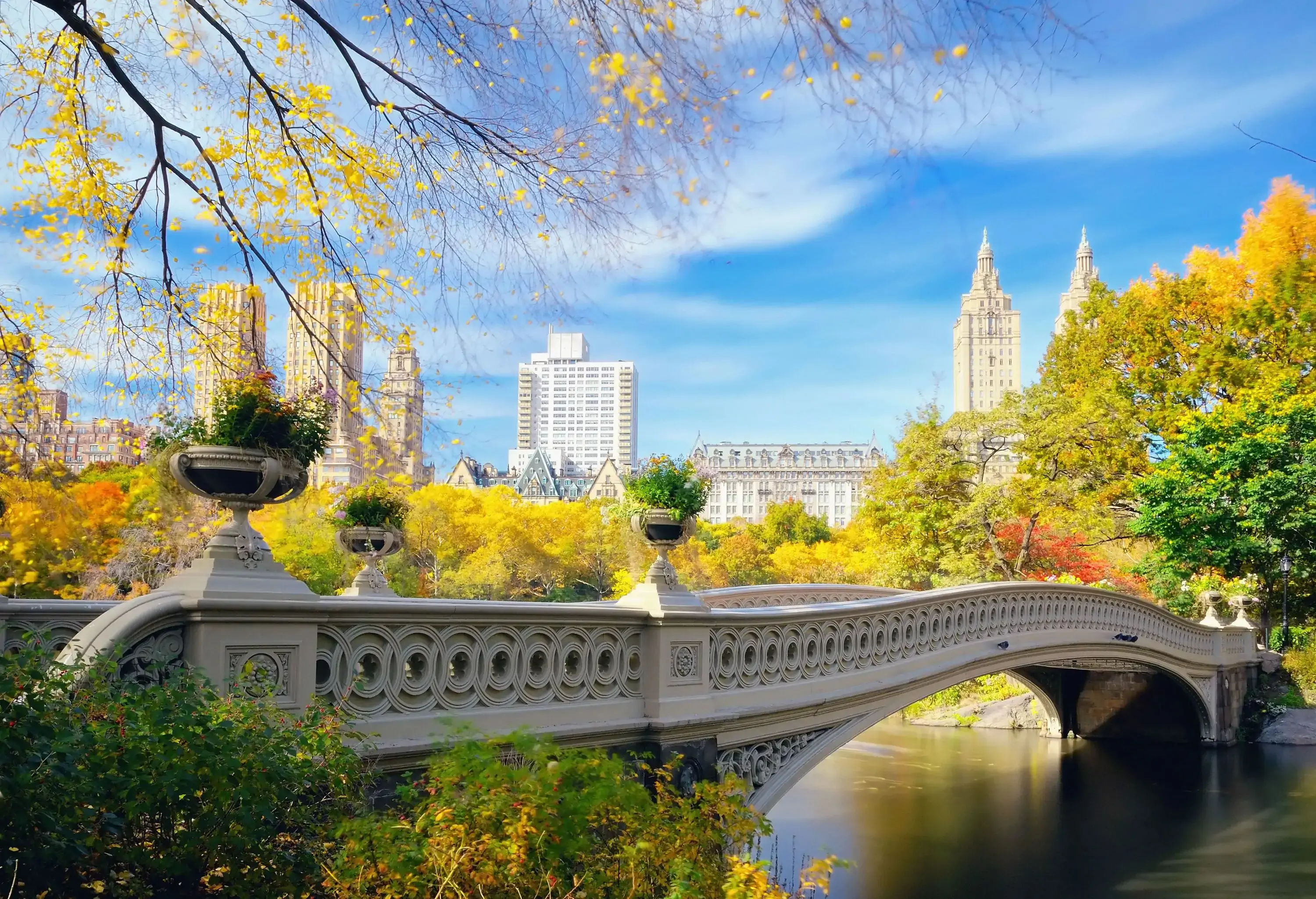 A cast iron bridge with planting urns across a calm lake surrounded by vibrant fall foliage.