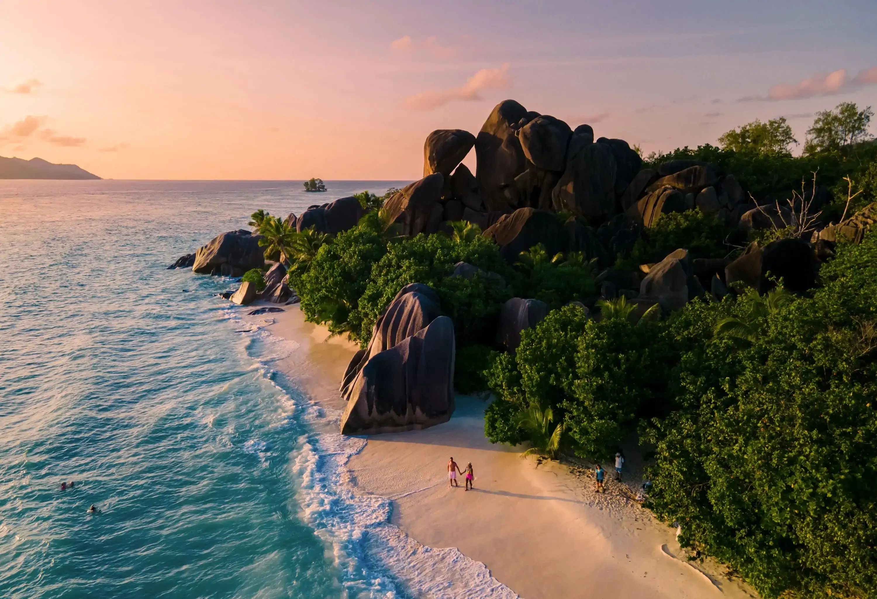 A naturally landscaped beach with rocks and lush trees visited by a couple and several friends on a scenic sunset.