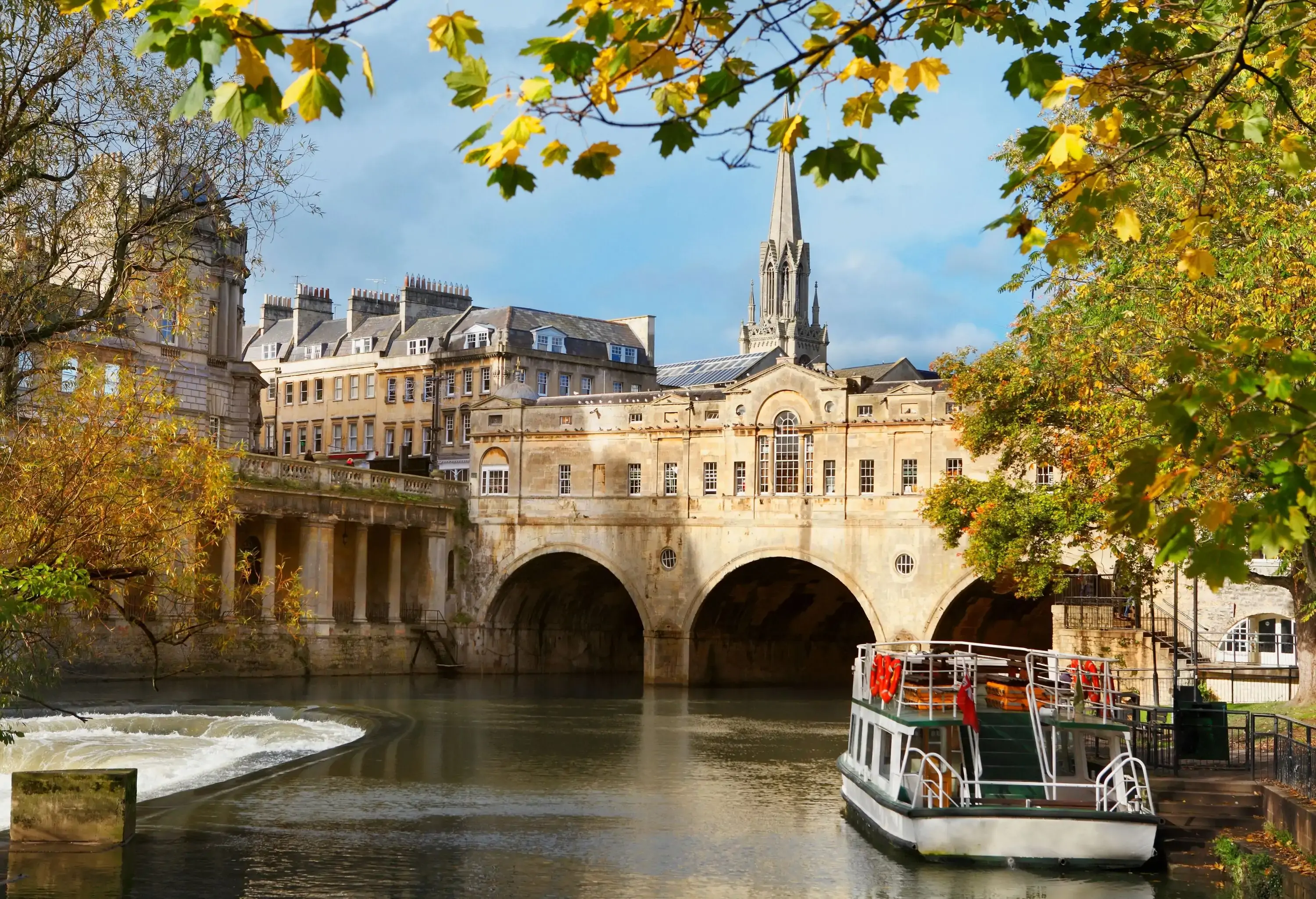 A boat close to a river dam is in front of an arch bridge with buildings on top.