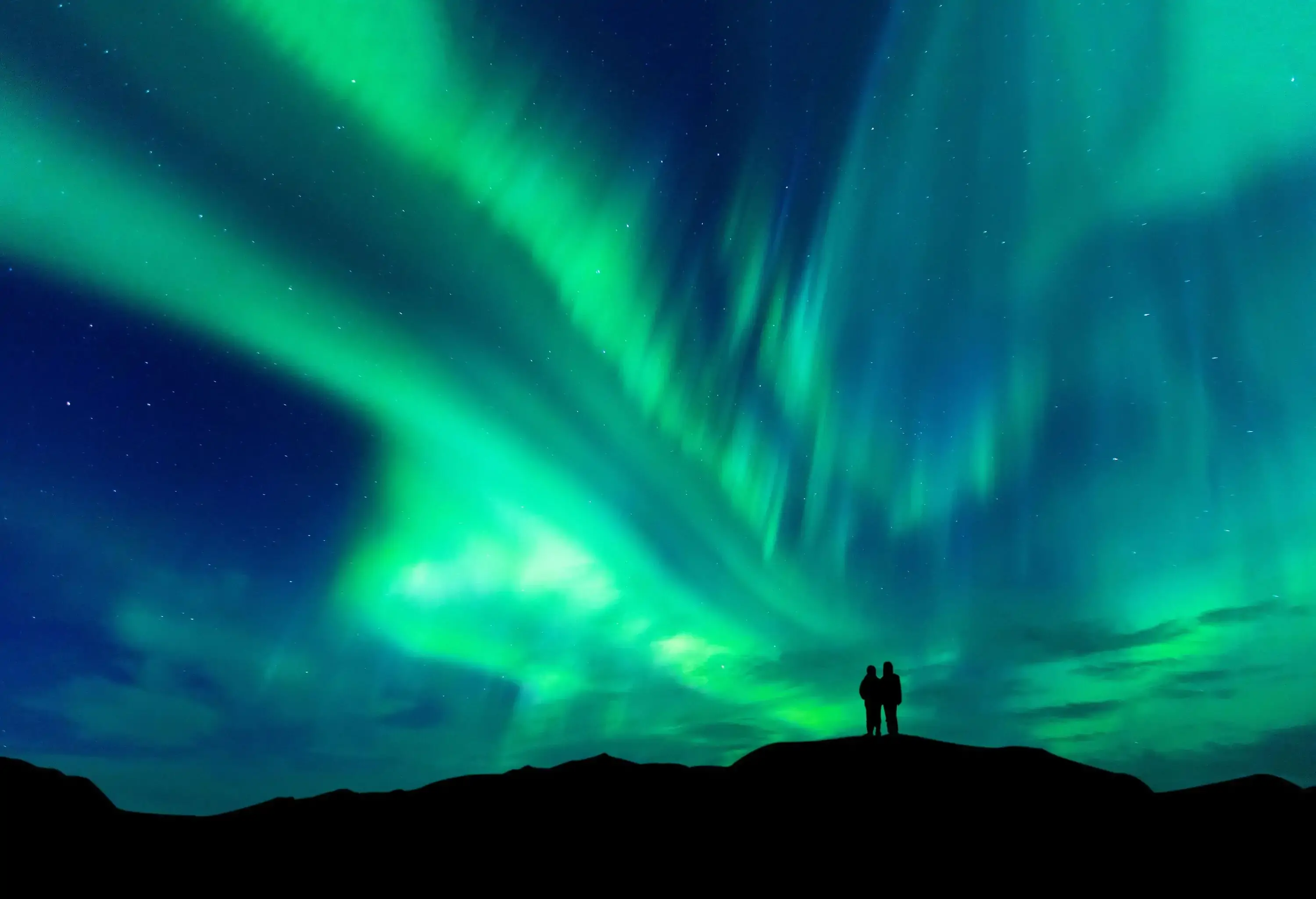 A shadow of two people standing on a mountain under the dazzling northern lights.