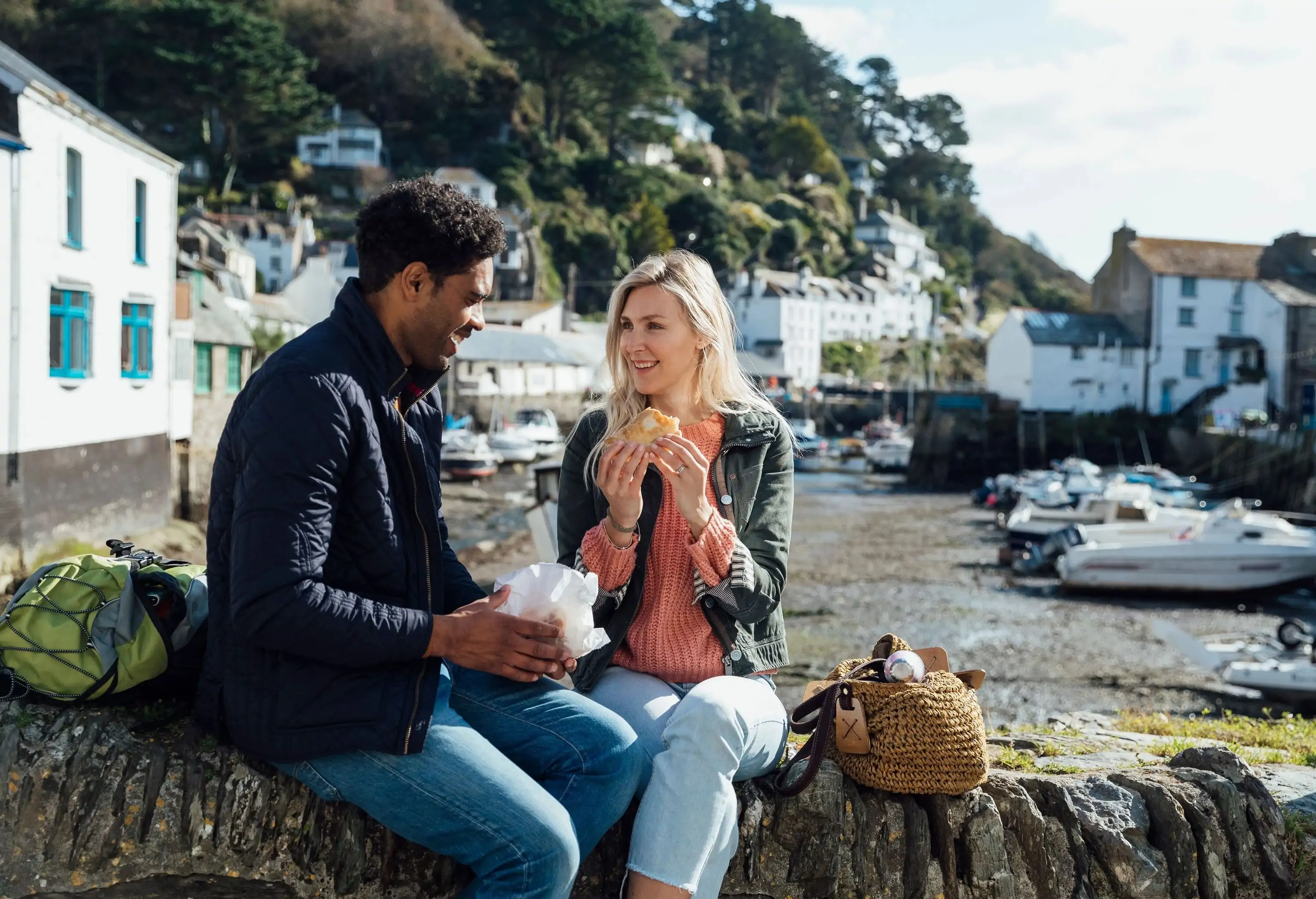 A couple eating and talking while seated on a rock wall with a view of white houses on the mountainside in the background.