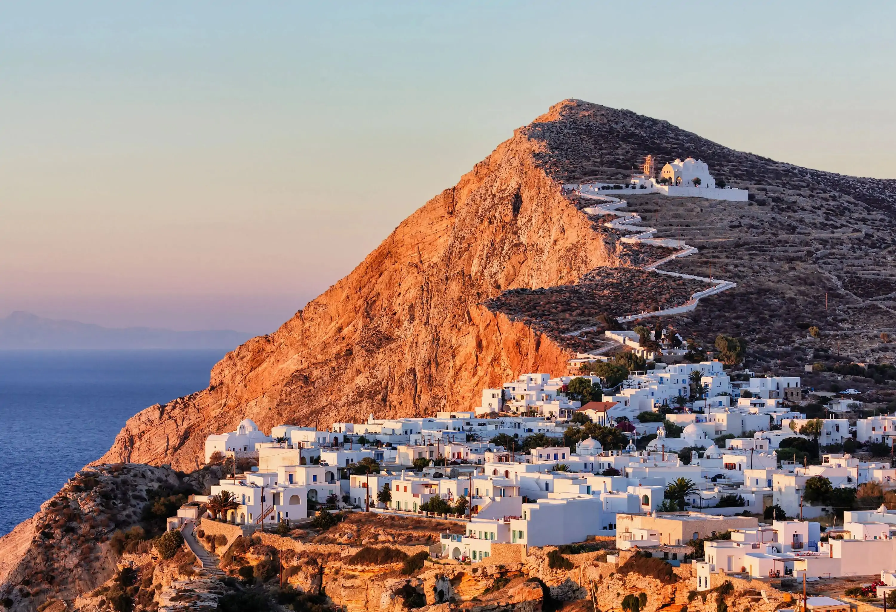 A white church situated at the top of a hill, with a winding road leading up to a white-washed hillside village that overlooks the sea.