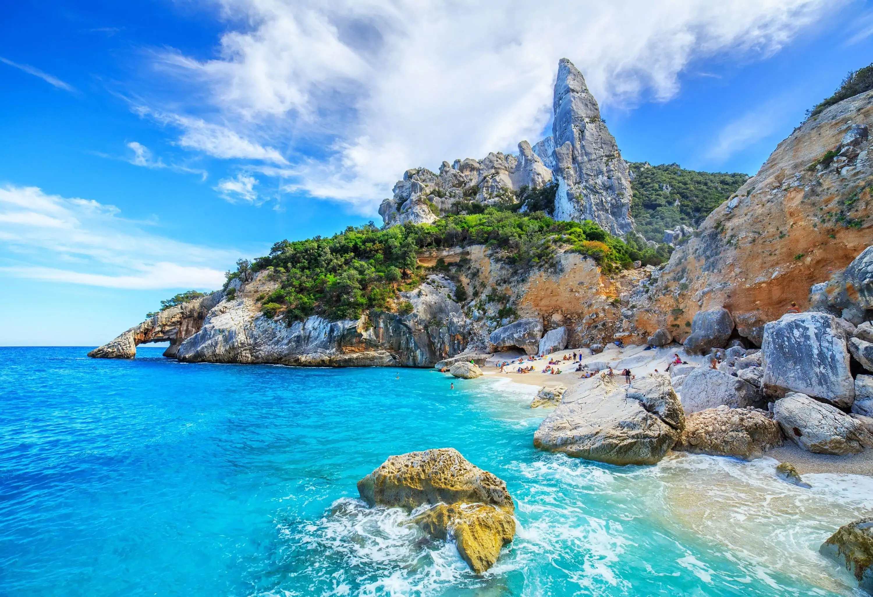 A small white sand beach with turquoise waters along boulders and a cliff topped with an imposing pointed rock.