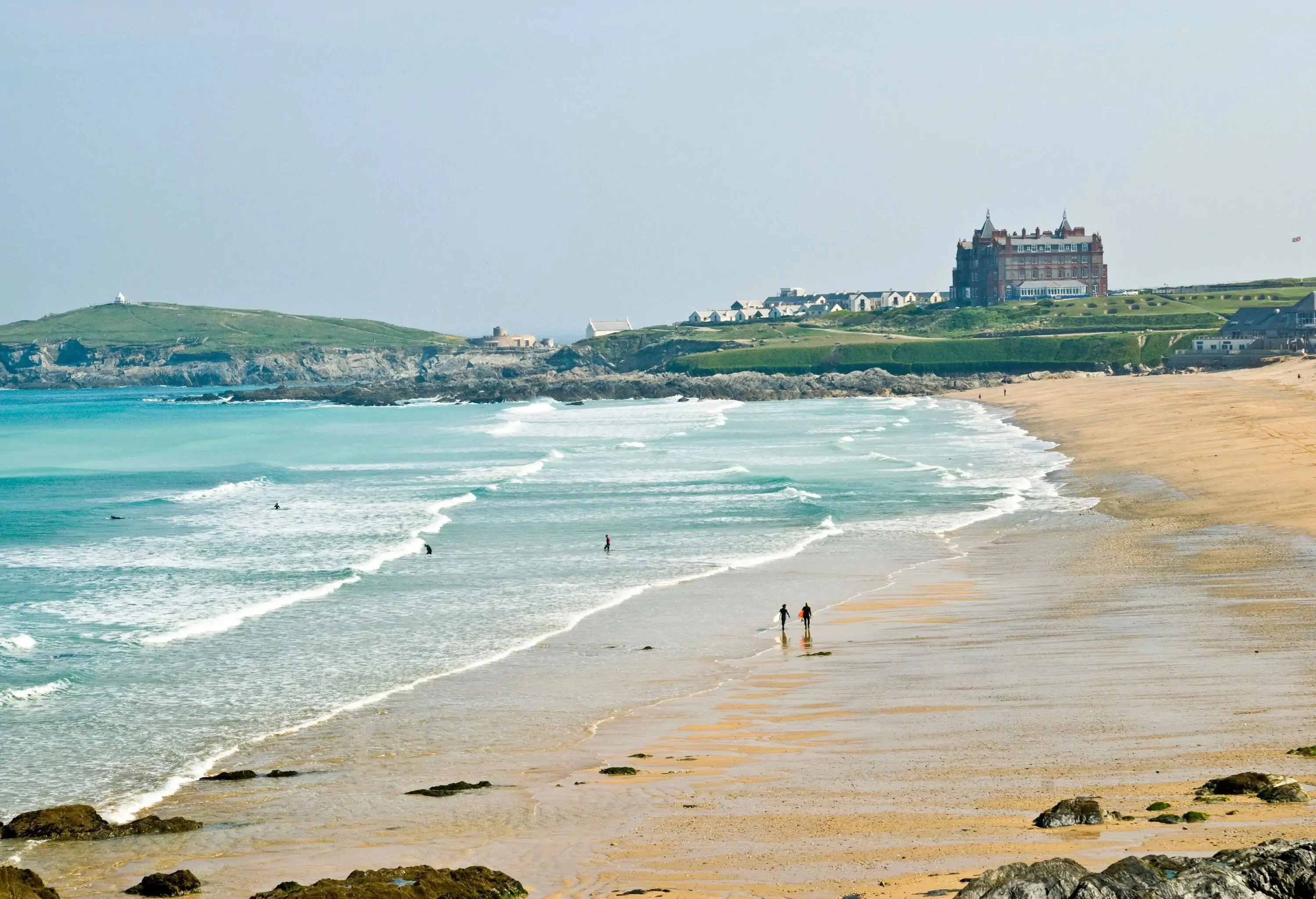 Surfers stroll down a beach with distant views of village houses and a huge building set on a hill.