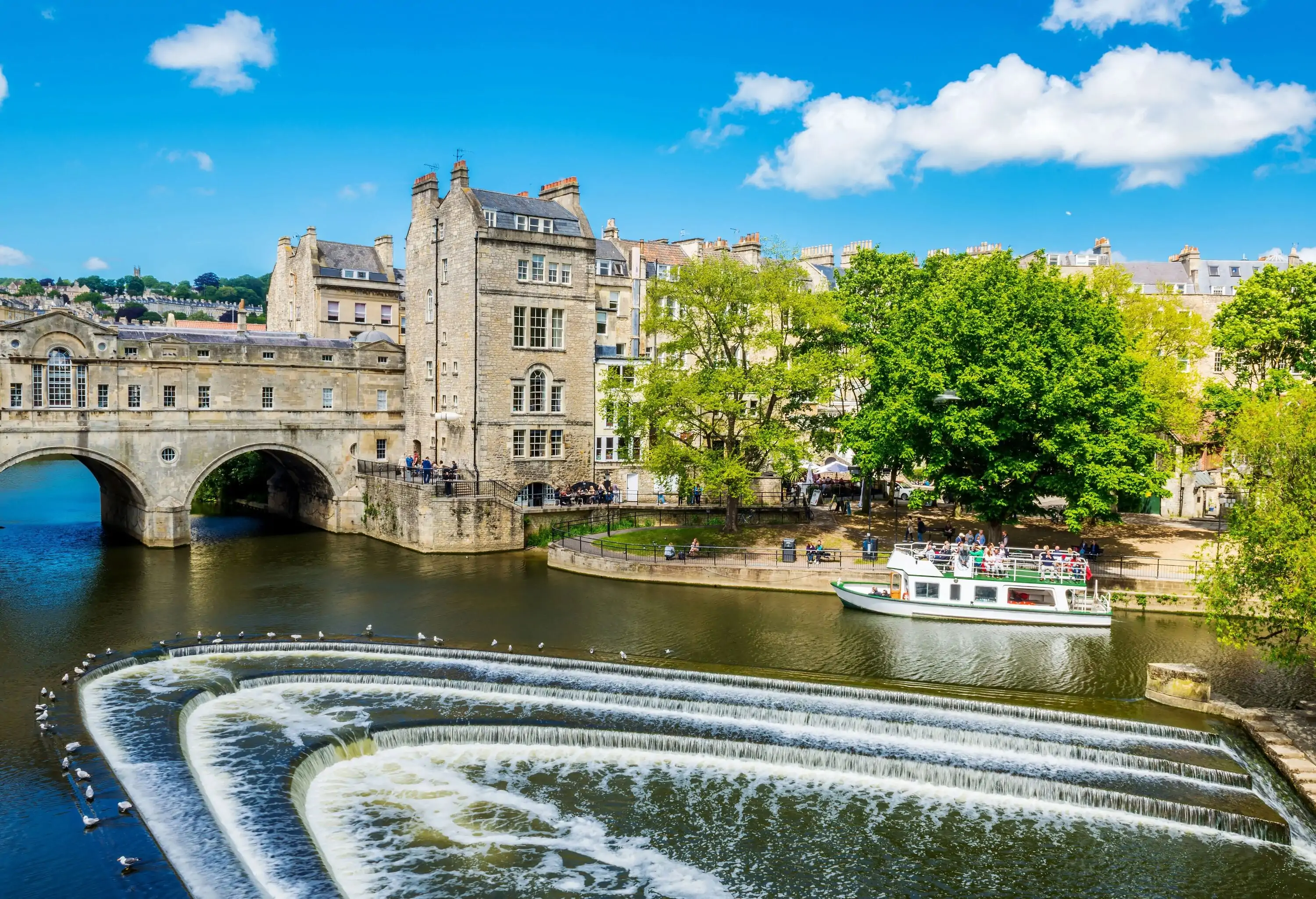 An arch bridge across a river with flocks of white birds standing at the edge of the weir and a small boat docked alongside a narrow walkway.