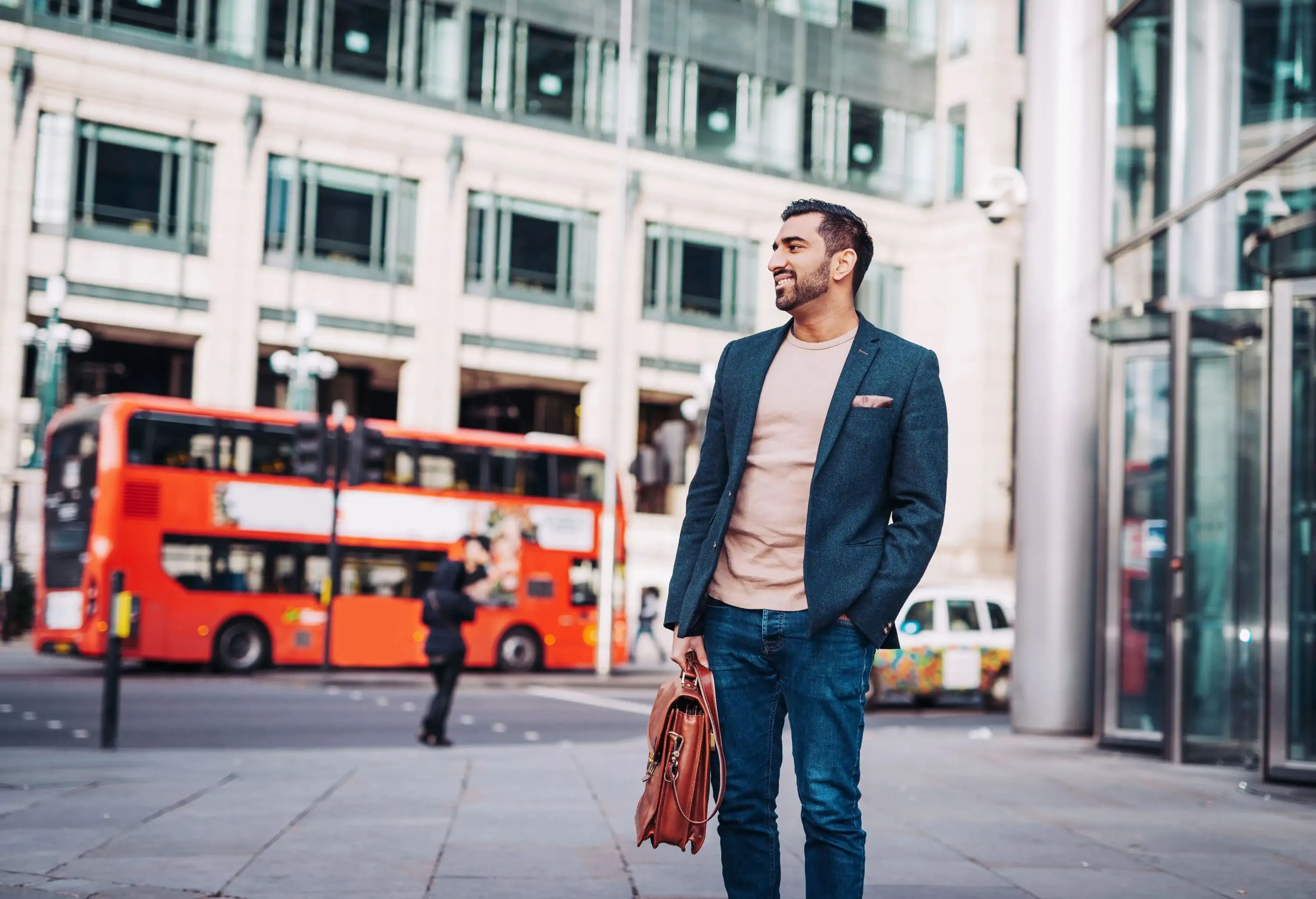 A happy man in smart casual attire stands in the street against a double-decker red bus on the road.